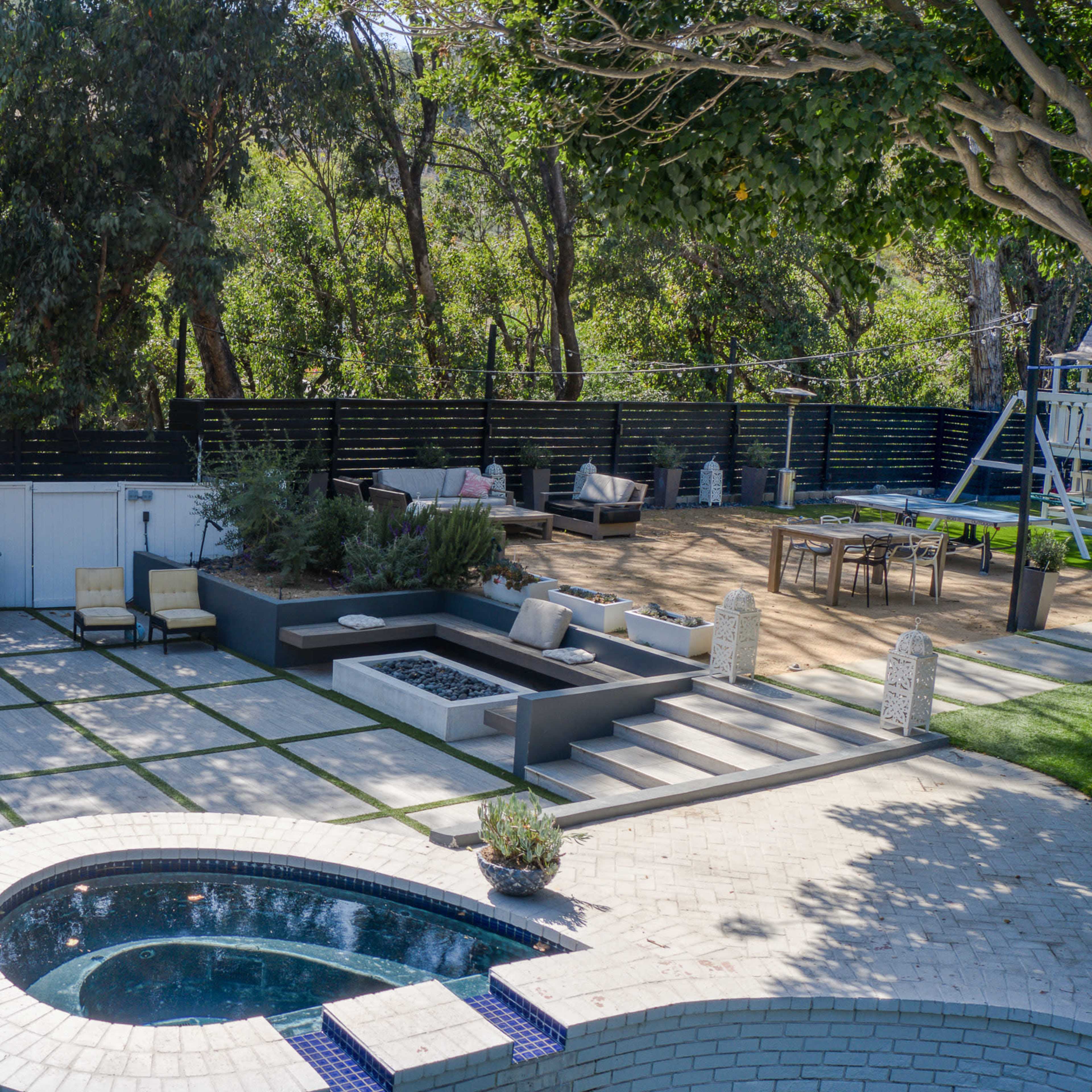 An outdoor patio area featuring a swimming pool, a bar with stools, a fire pit, and a lounge space surrounded by trees.