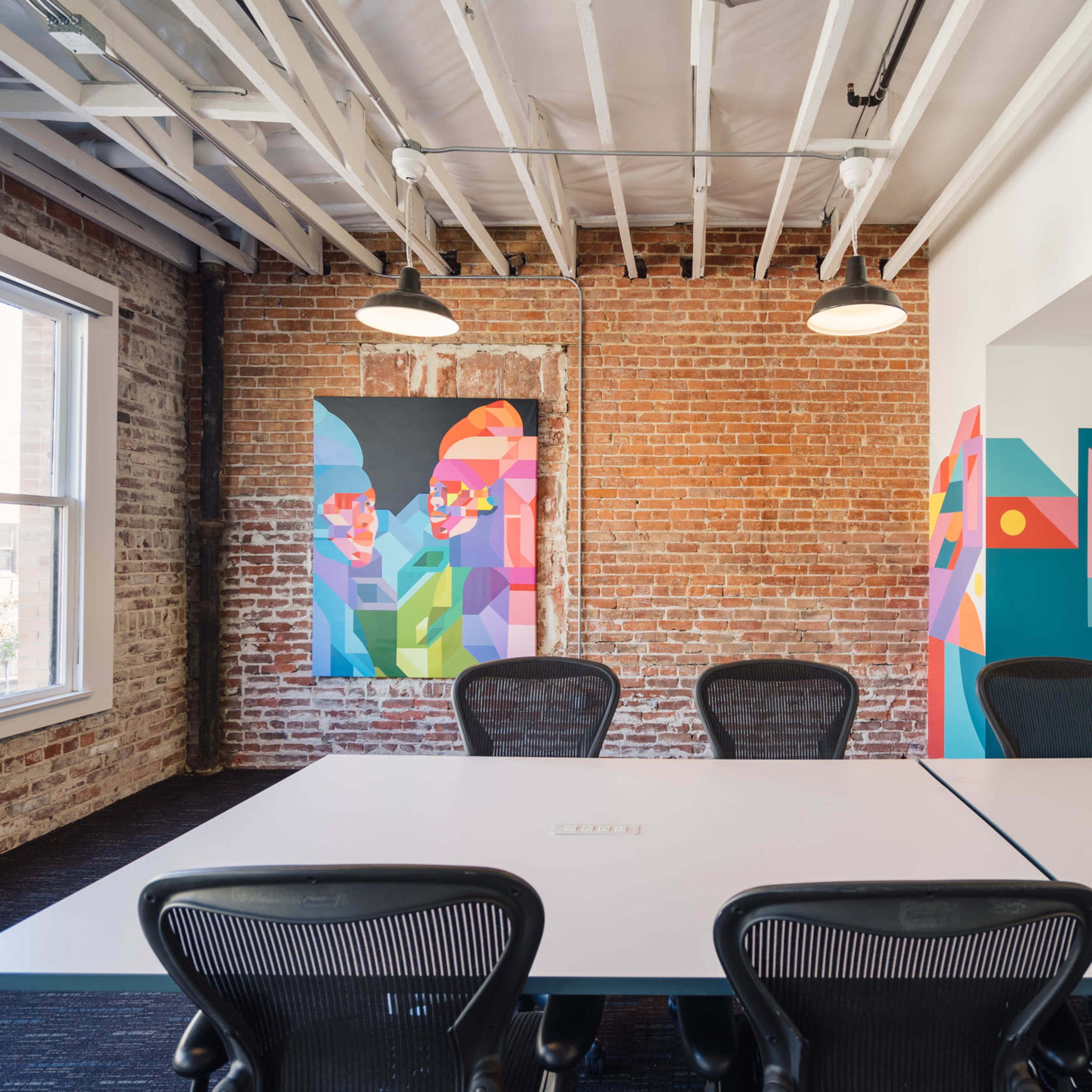 A modern office space with exposed brick walls, a long white table, and colorful abstract artwork on the walls.