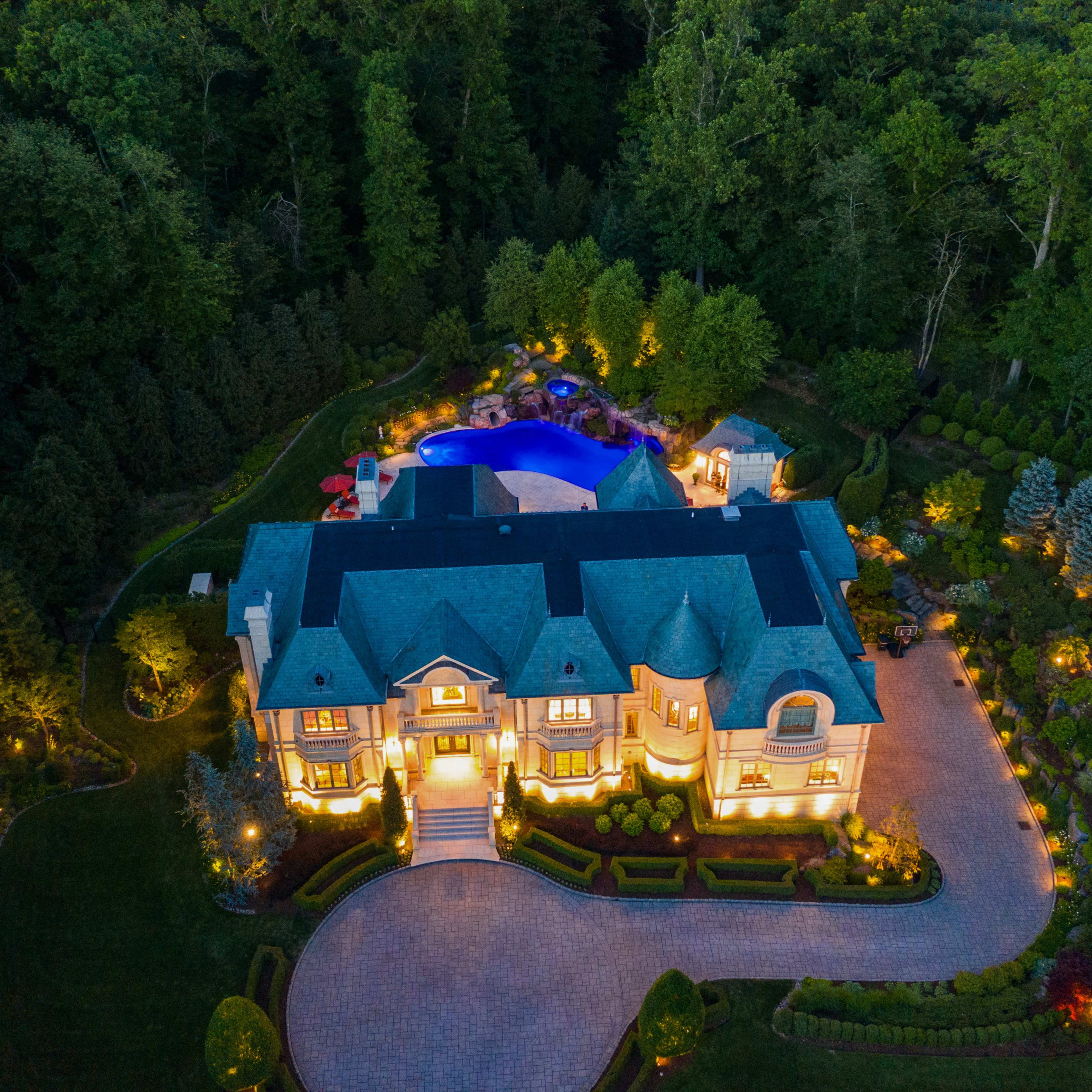 An aerial view of a large, illuminated mansion surrounded by lush greenery and a backyard pool area.