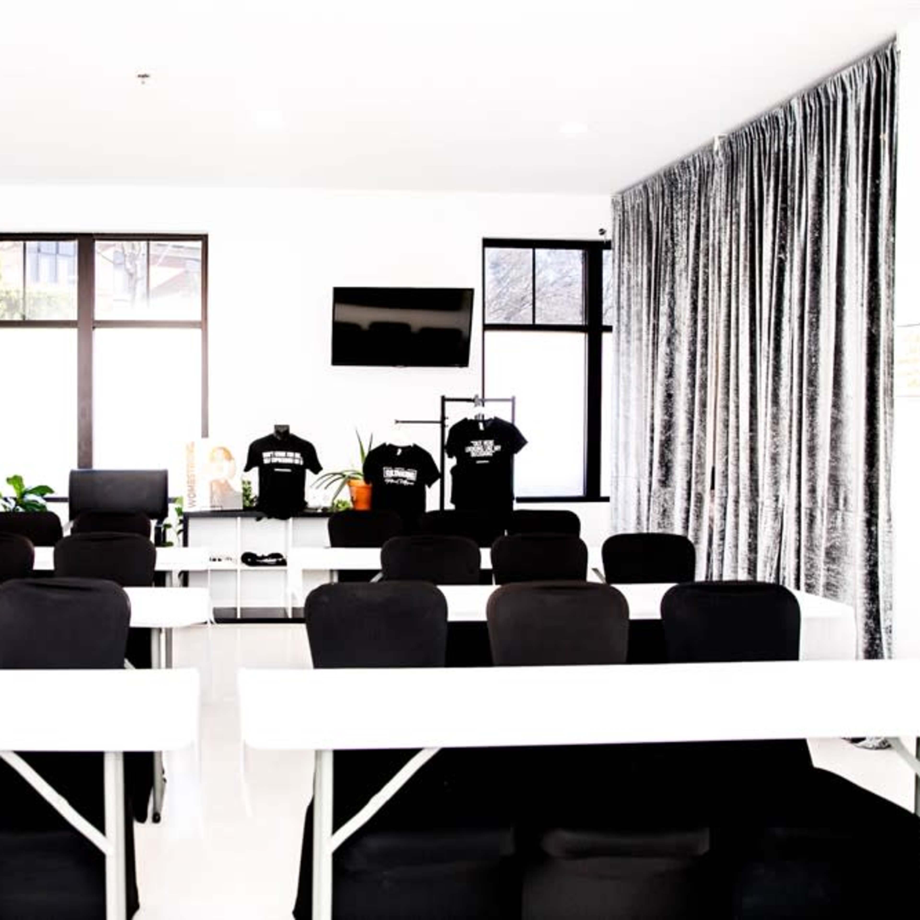 A modern meeting room features rows of black chairs facing a white table, with large windows and a television on the wall.