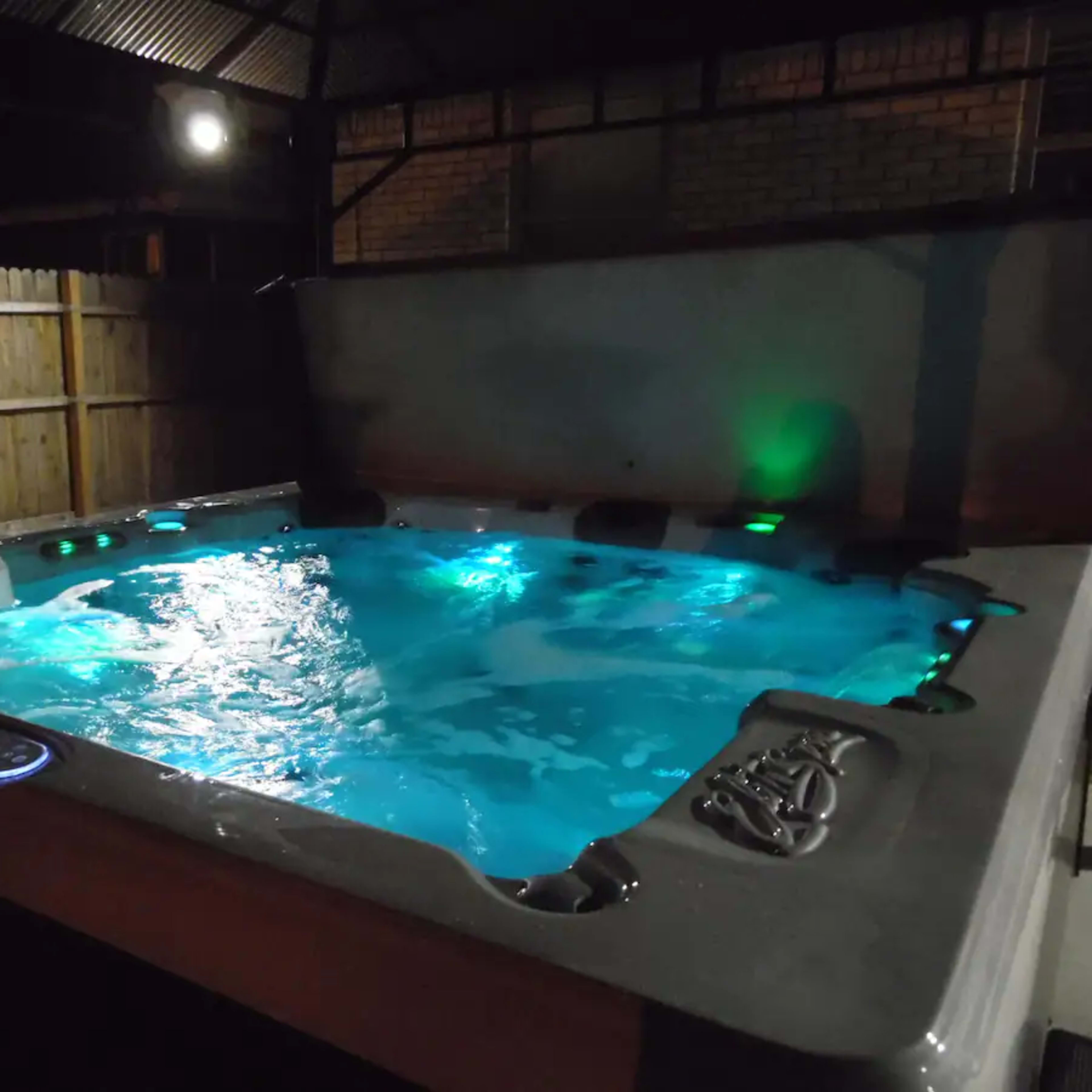 A hot tub with illuminated water is situated under a covered outdoor area at night.