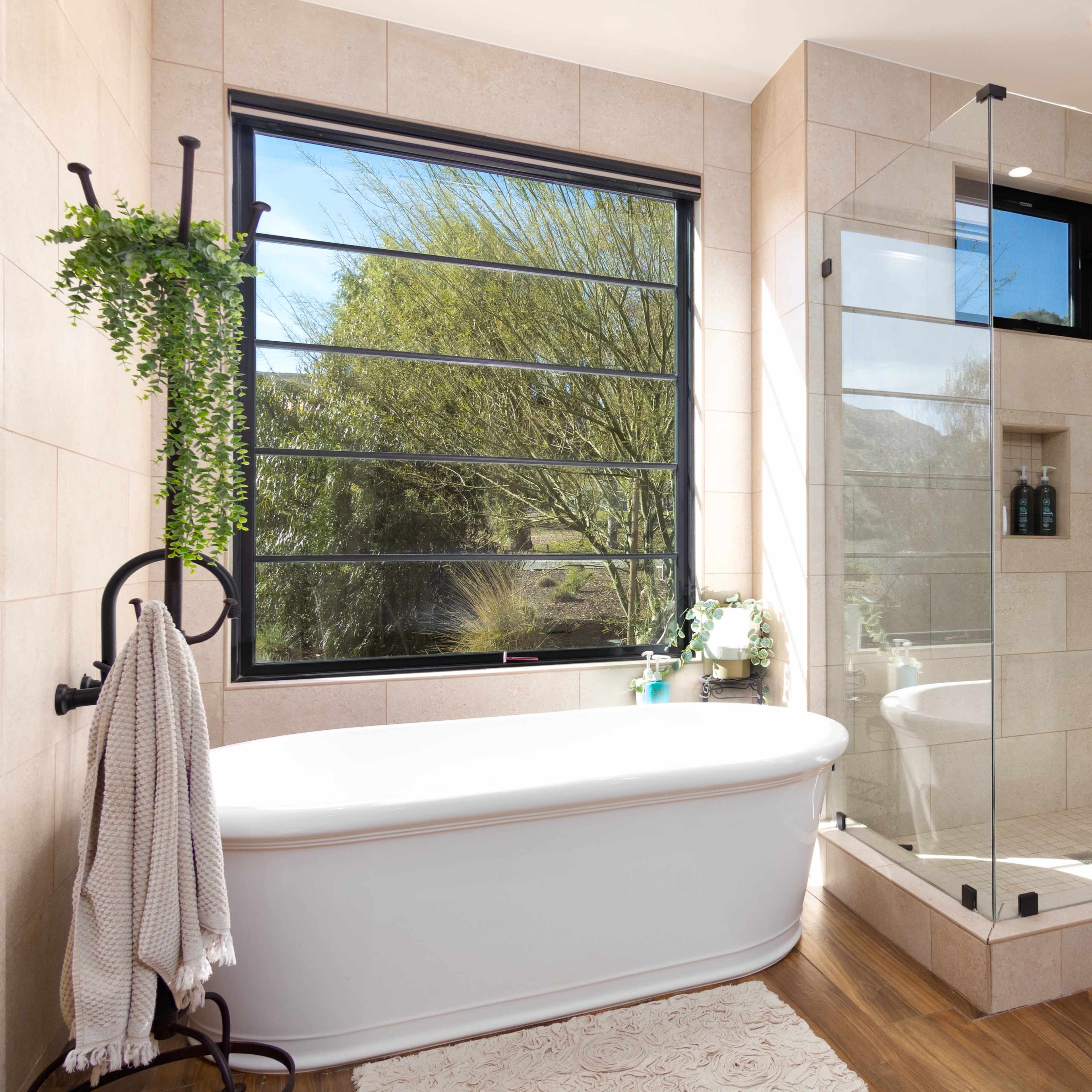 The bathroom features a freestanding tub next to a glass-enclosed shower with a textured wood wall and a window showcasing a view of greenery outside.