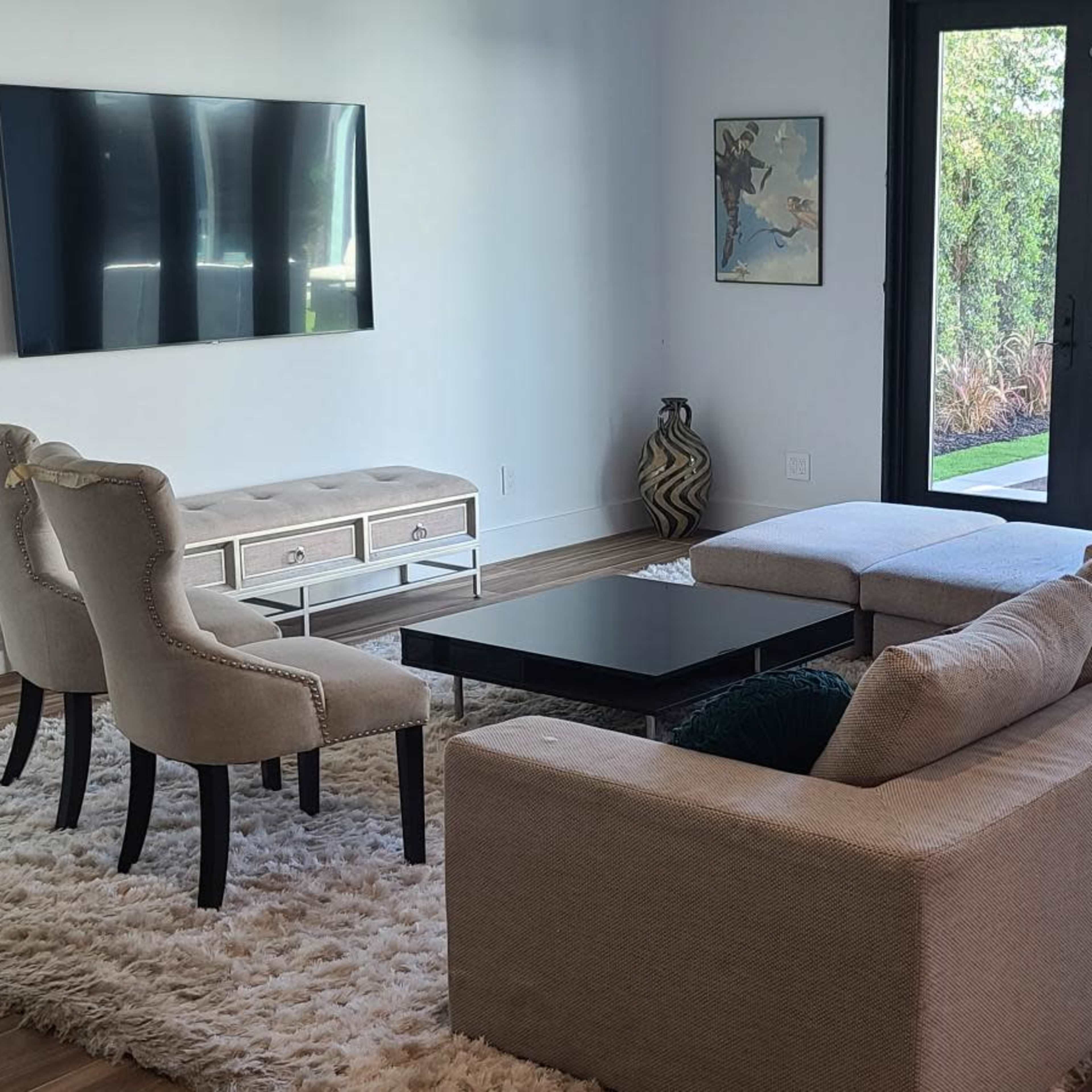 A modern living room with a sectional sofa, coffee table, and a wall-mounted television, featuring large windows that provide a view of an outdoor space.