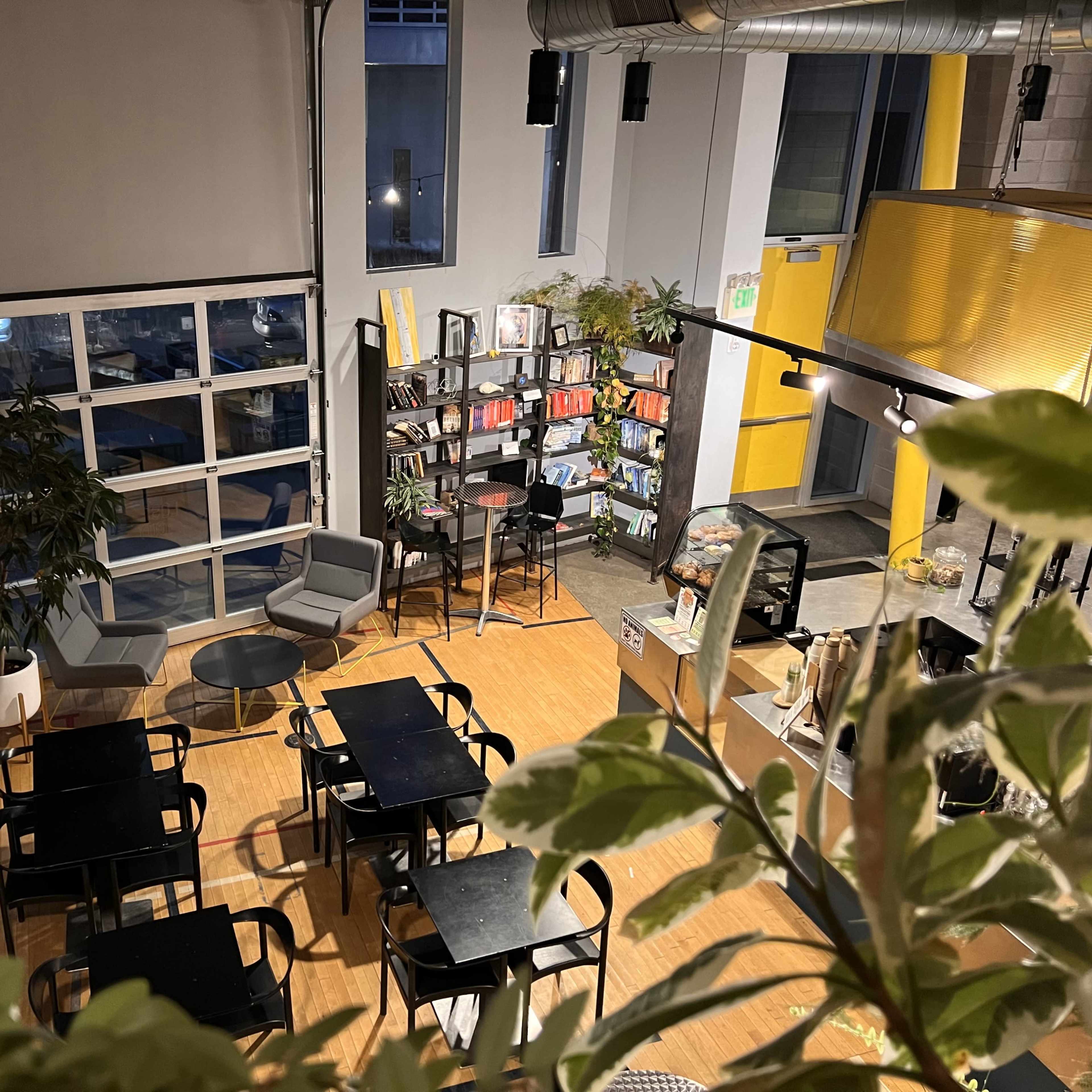 A modern café with several black tables and chairs, bookshelves, and large windows allowing natural light to enter.