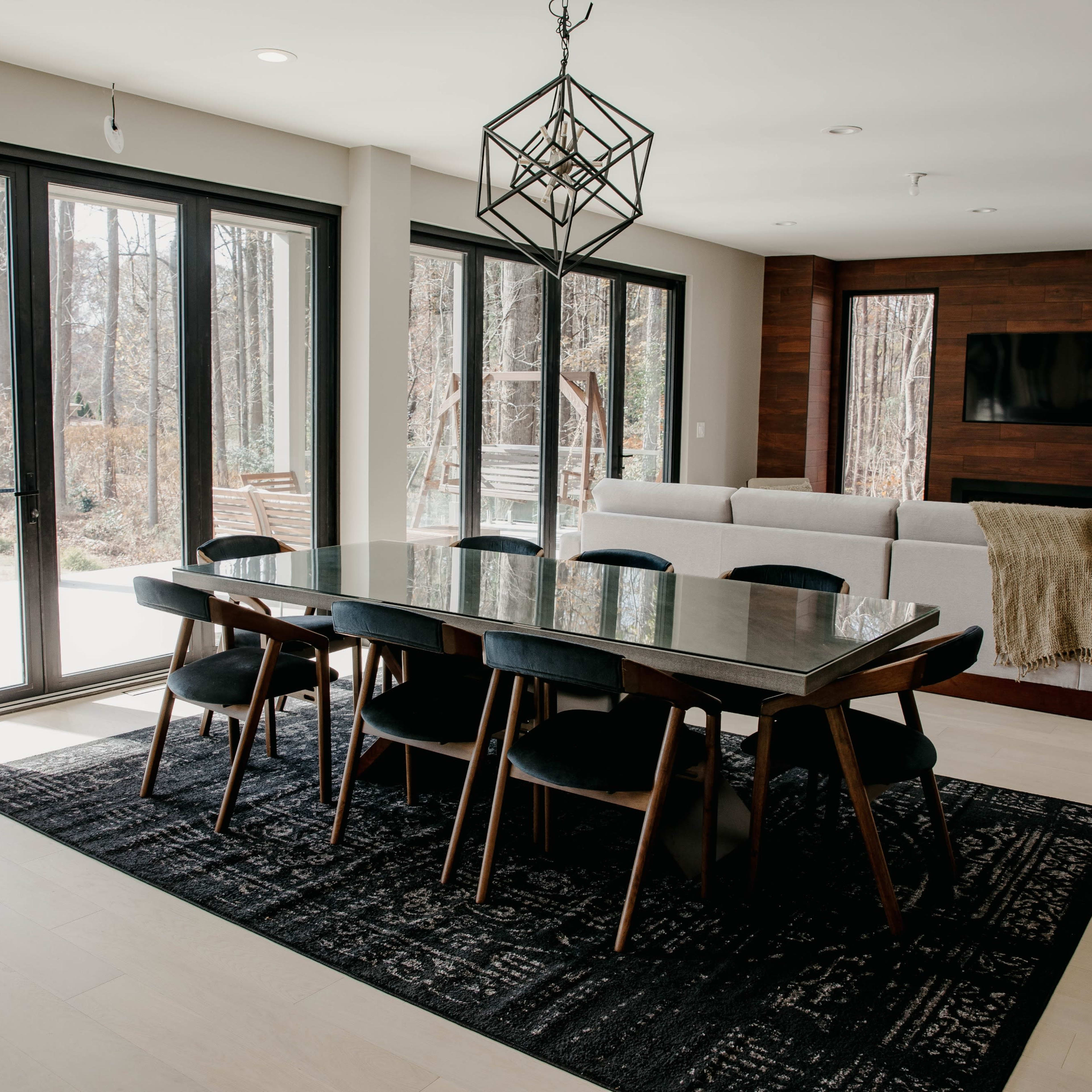 A modern dining area features a rectangular table with black chairs, surrounded by large glass doors that open to a wooded view.