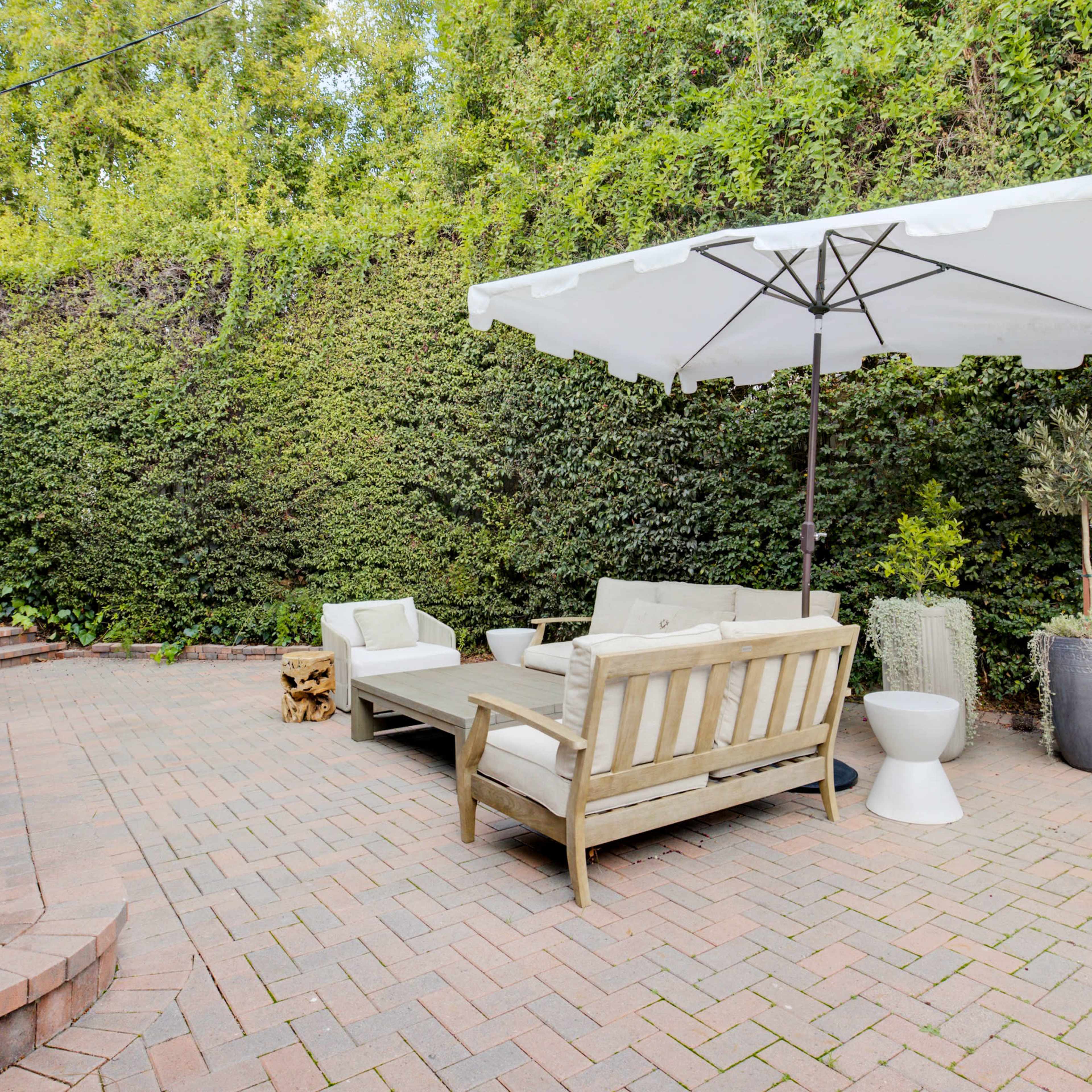 A patio area featuring a wooden seating arrangement, a large umbrella, and potted plants surrounded by a lush green wall.