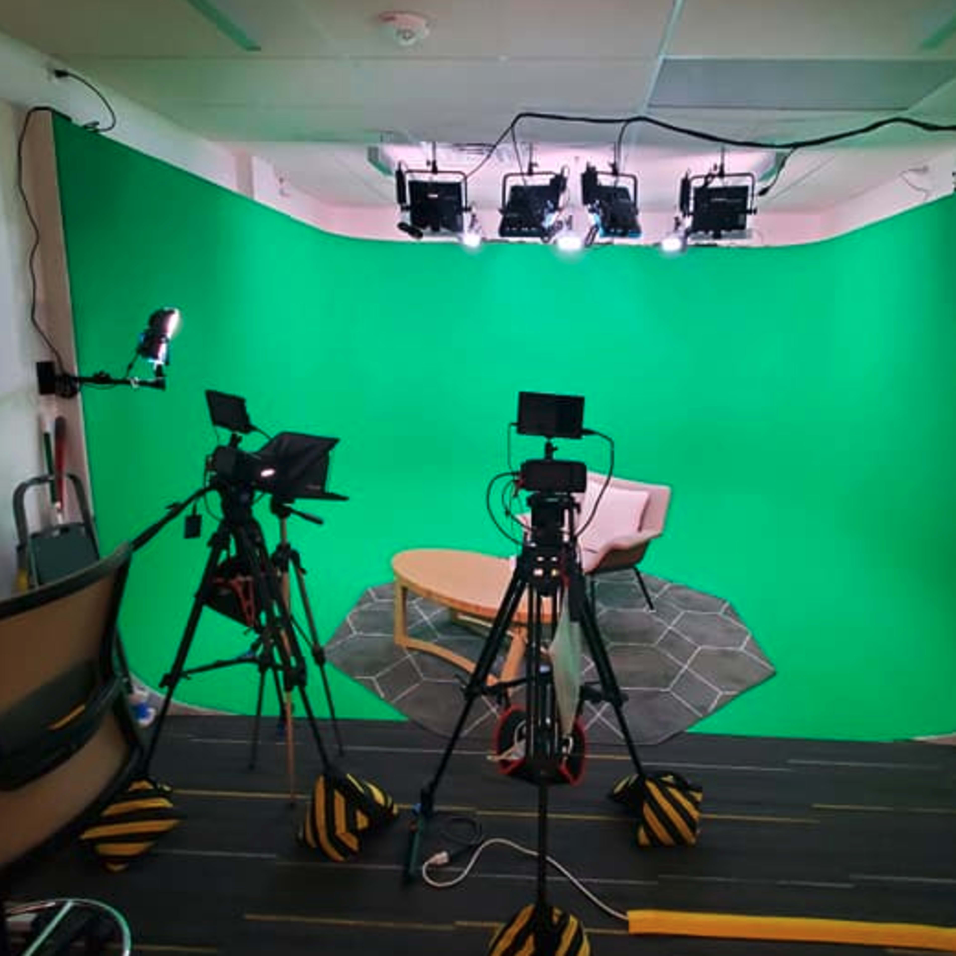 A video recording studio set up with two tripods holding cameras, a green backdrop, and overhead lights illuminating a small seating area with a beige chair and a coffee table.