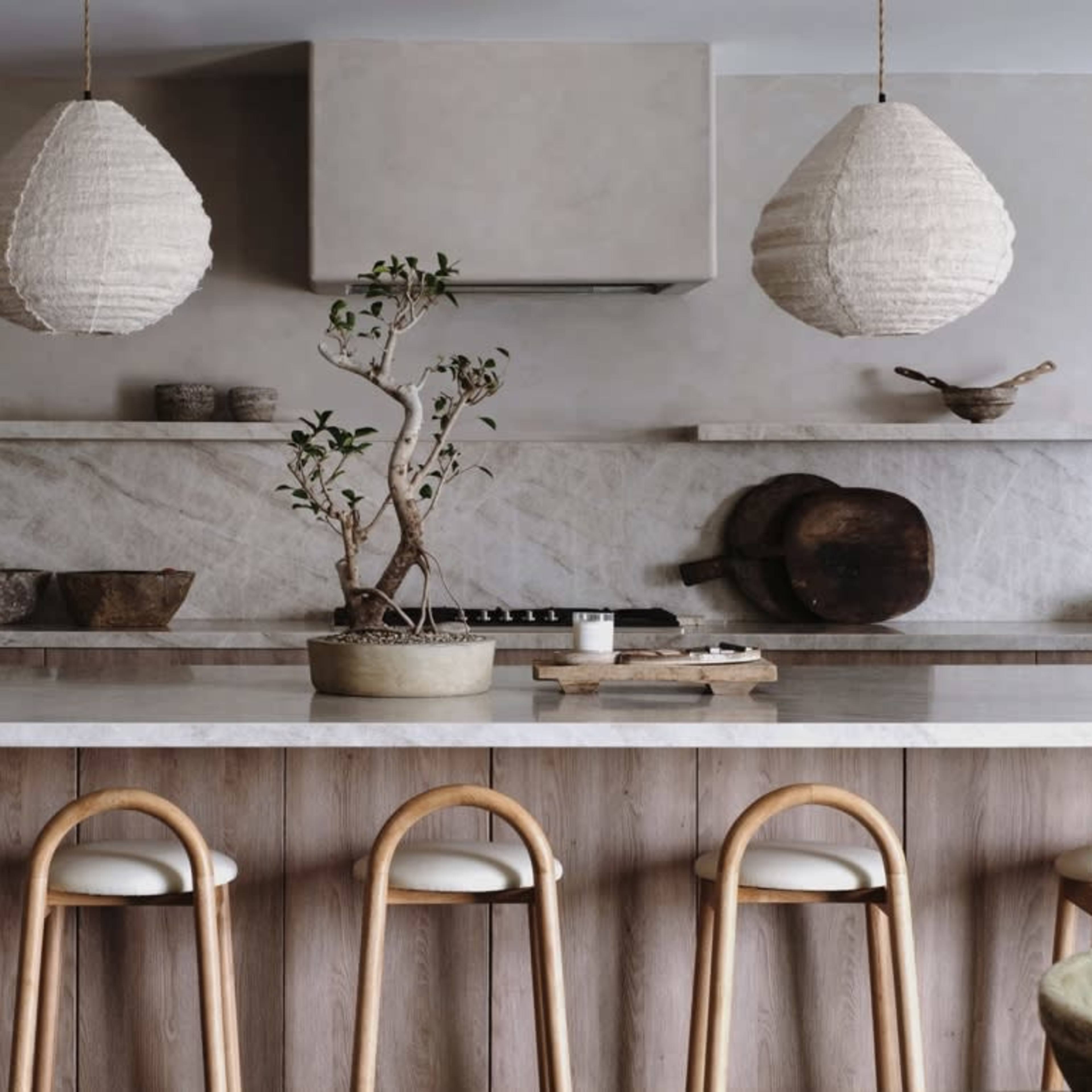 A modern kitchen with a large marble island, wooden bar stools, and pendant lighting, featuring a minimalist design with natural elements.