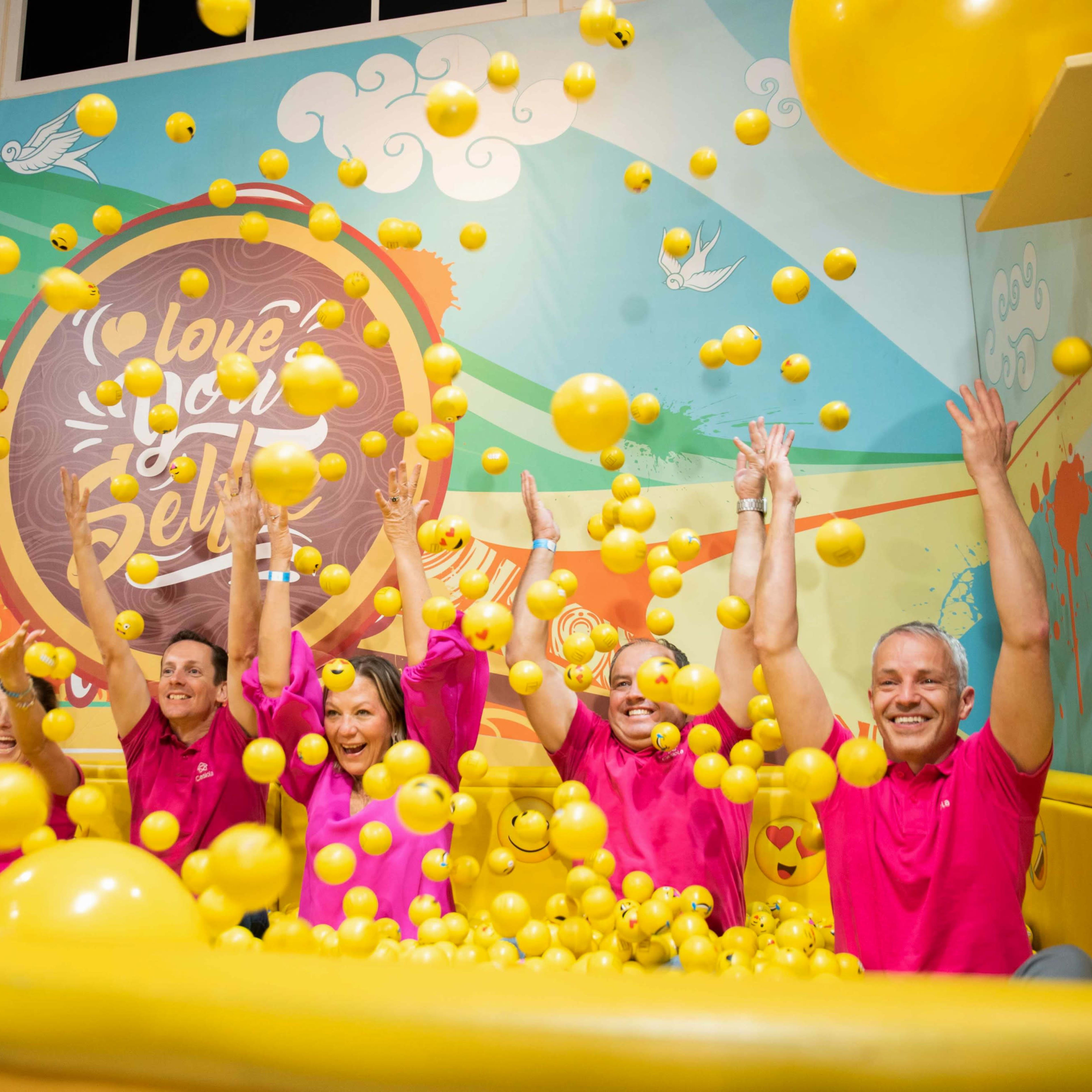 A group of six people in pink shirts joyfully toss yellow balls in a colorful indoor play area.