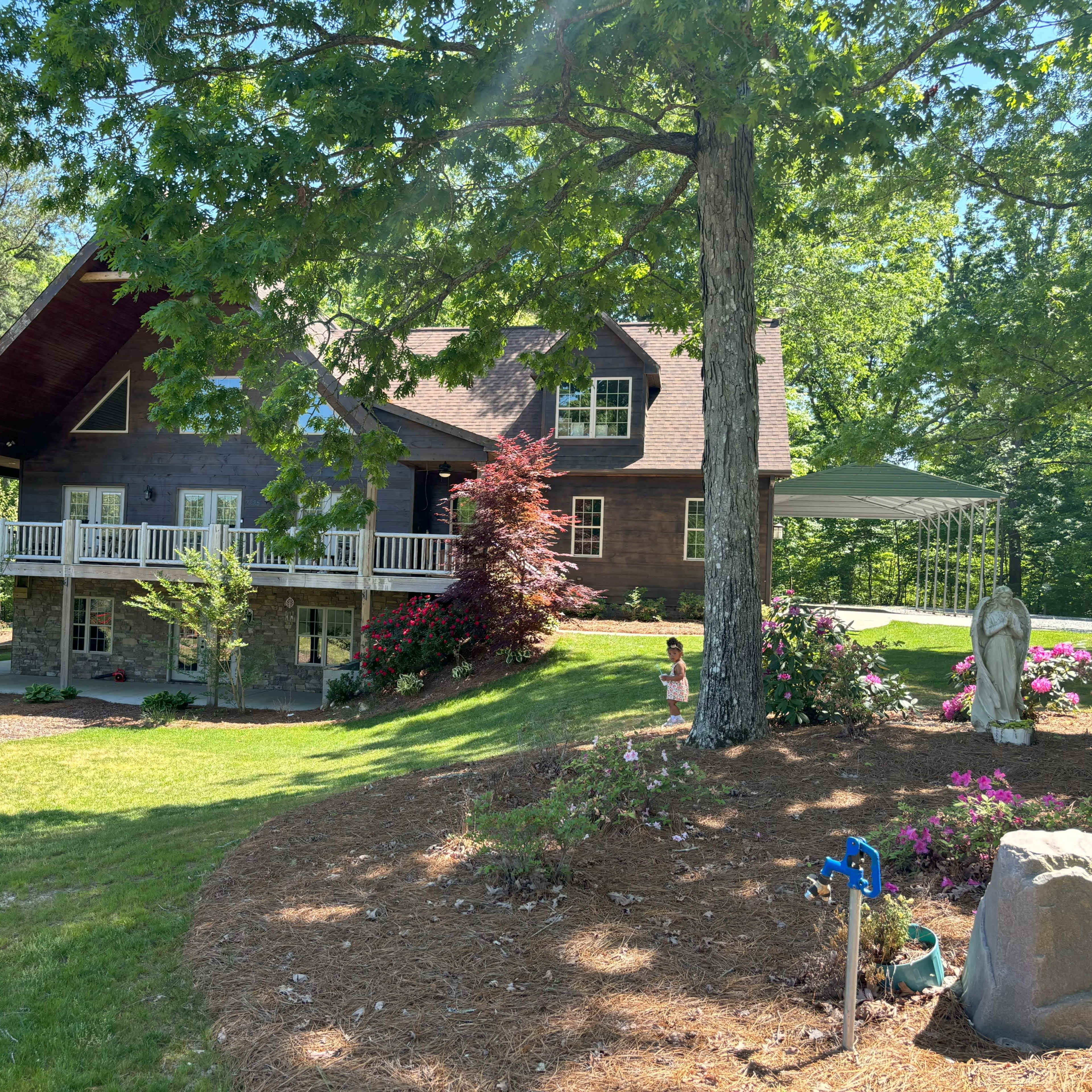 A large two-story wooden house with a porch is set in a green landscape surrounded by trees and flower beds.