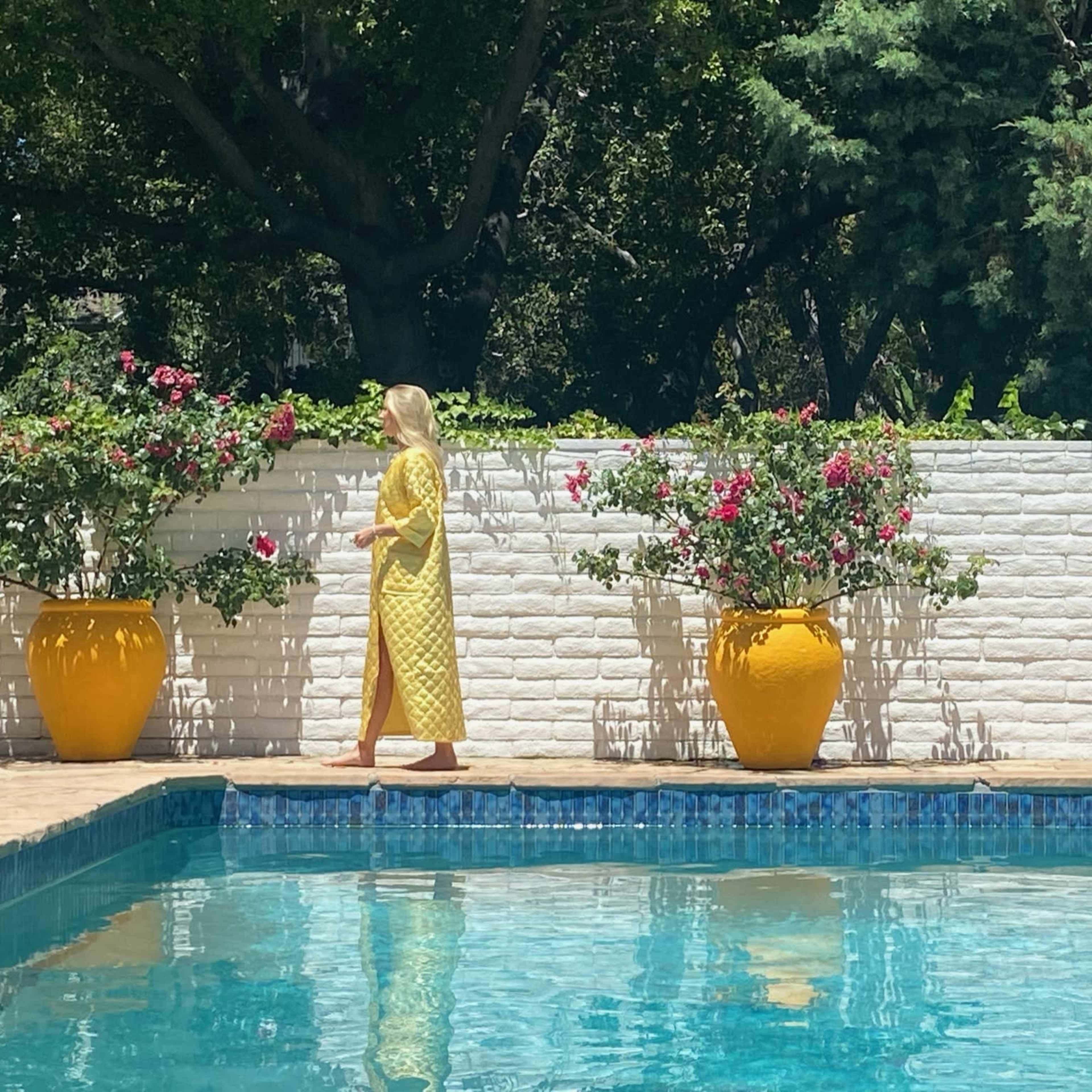 A woman in a yellow dress walks beside a swimming pool with yellow pots and blooming flowers against a white wall.