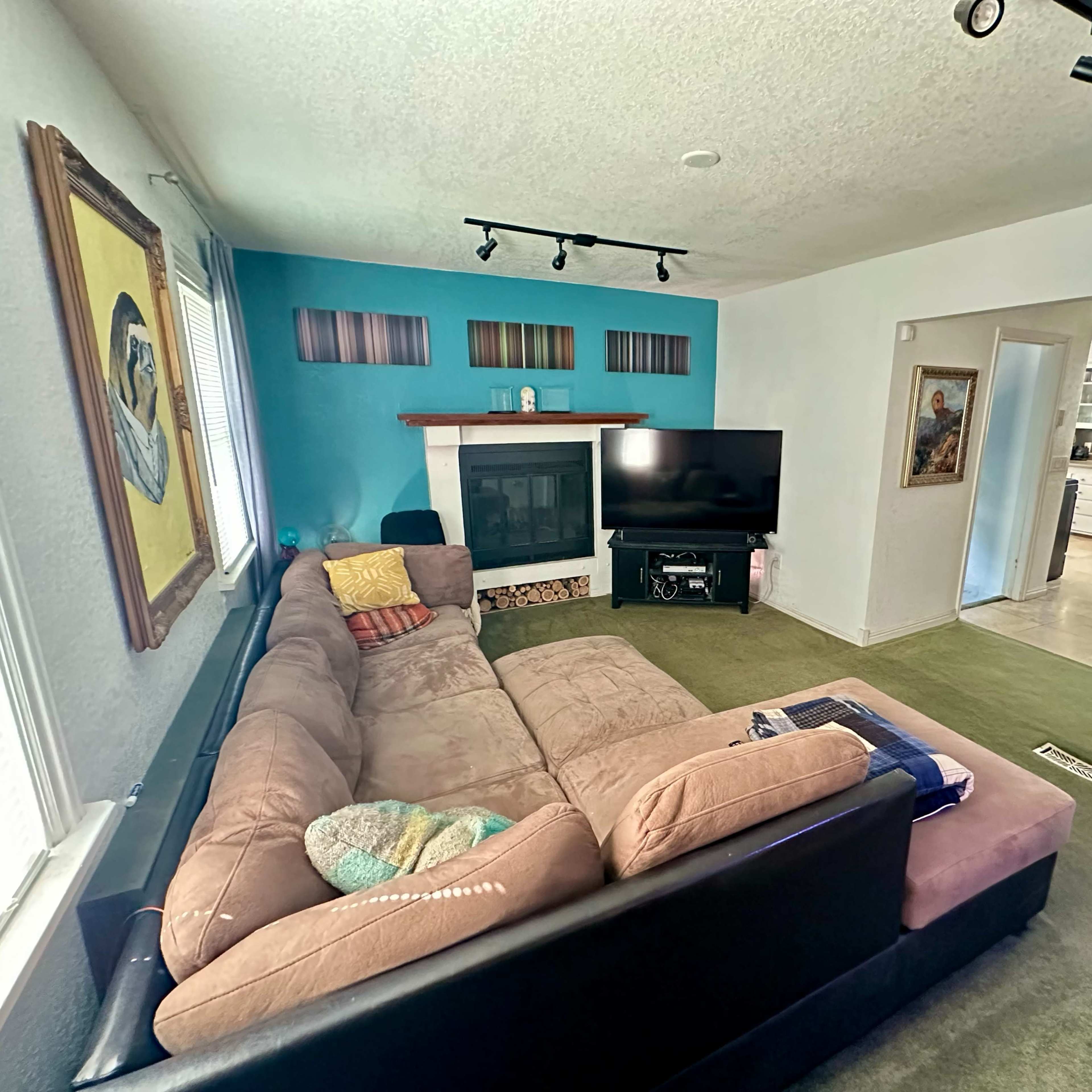 A living room with a sectional couch, a fireplace, a television, and a visible kitchen area in the background.