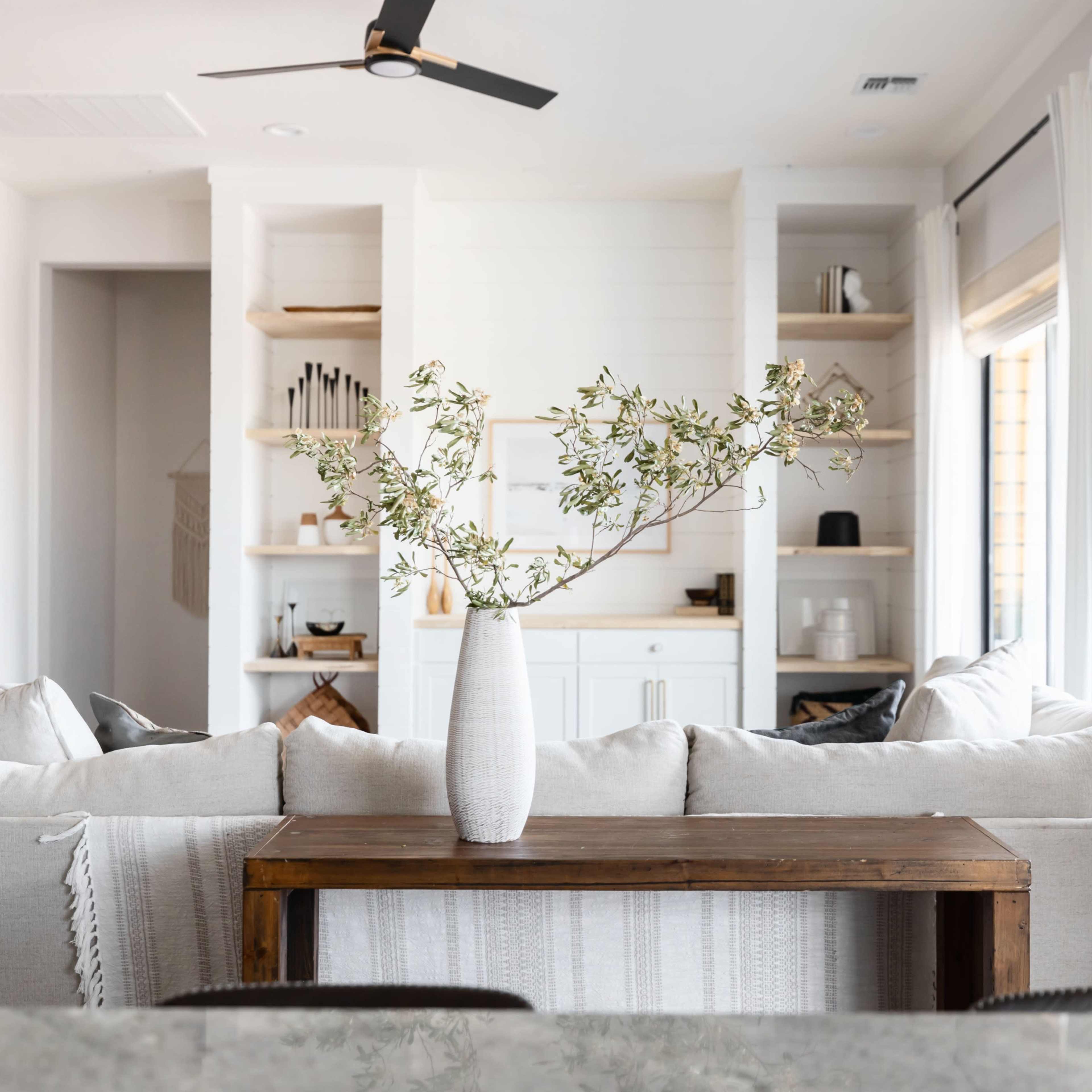 A light-filled living space features a beige sofa with a wooden table in front, adorned with a vase of greenery, and a backdrop of open shelves and windows.