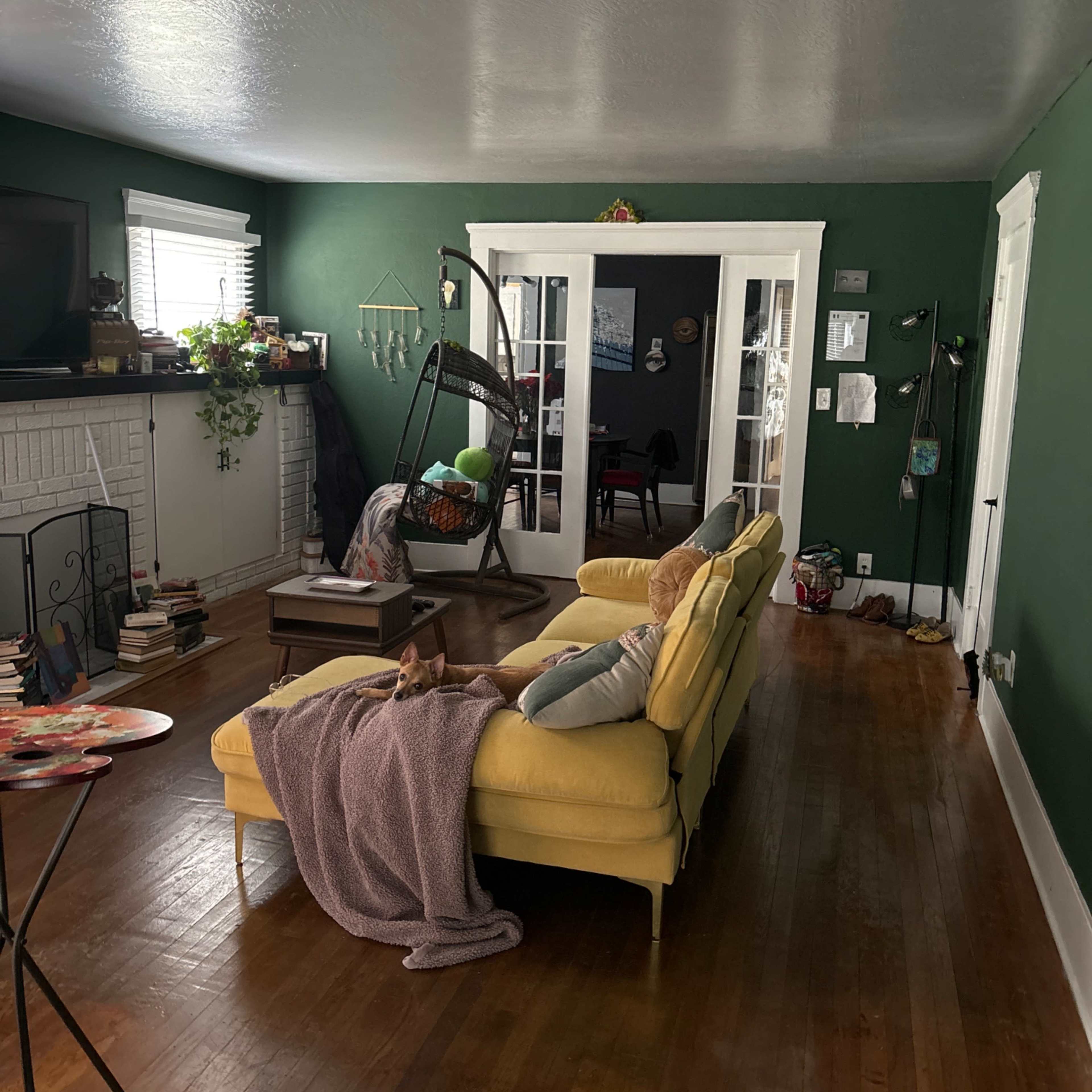 A cozy living room with a yellow sofa, a green accent wall, and wooden flooring, featuring a television and various decorative items.