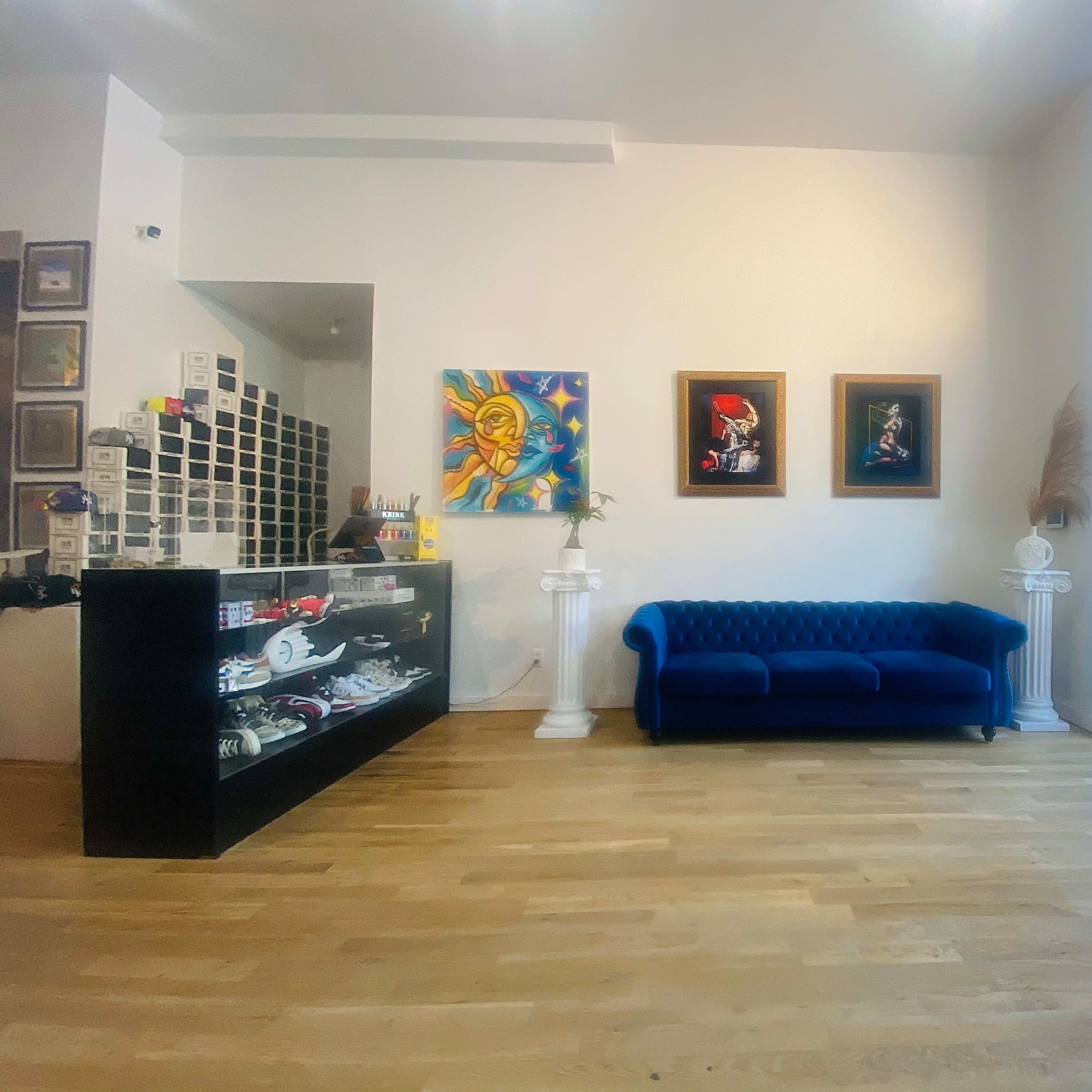 A modern interior space featuring a blue sofa, two framed artworks, and a glass display counter, all set against a light-colored wall and wooden floor.