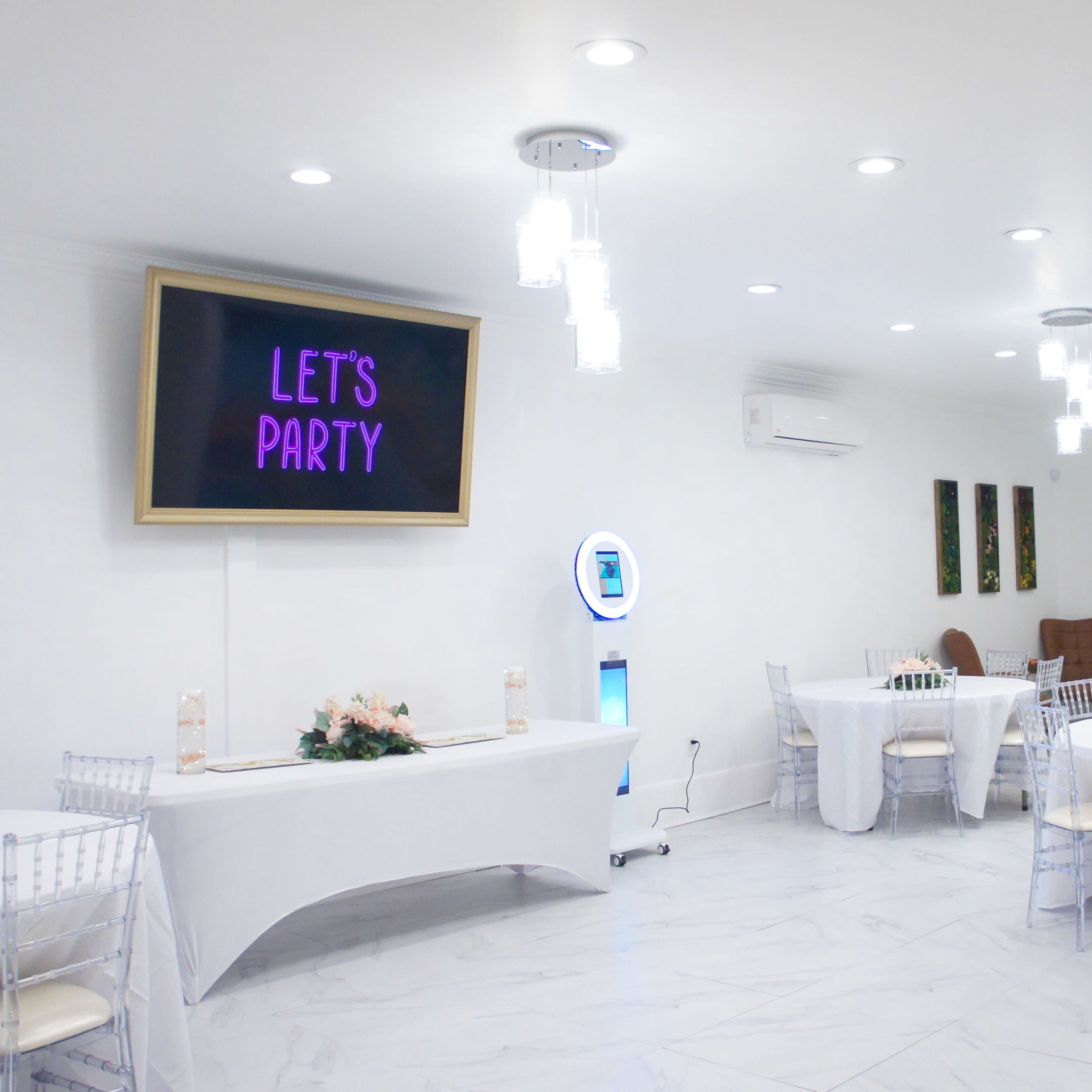 A bright, modern event space with white walls, round tables covered in white linens, and a screen displaying the text "LET'S PARTY."