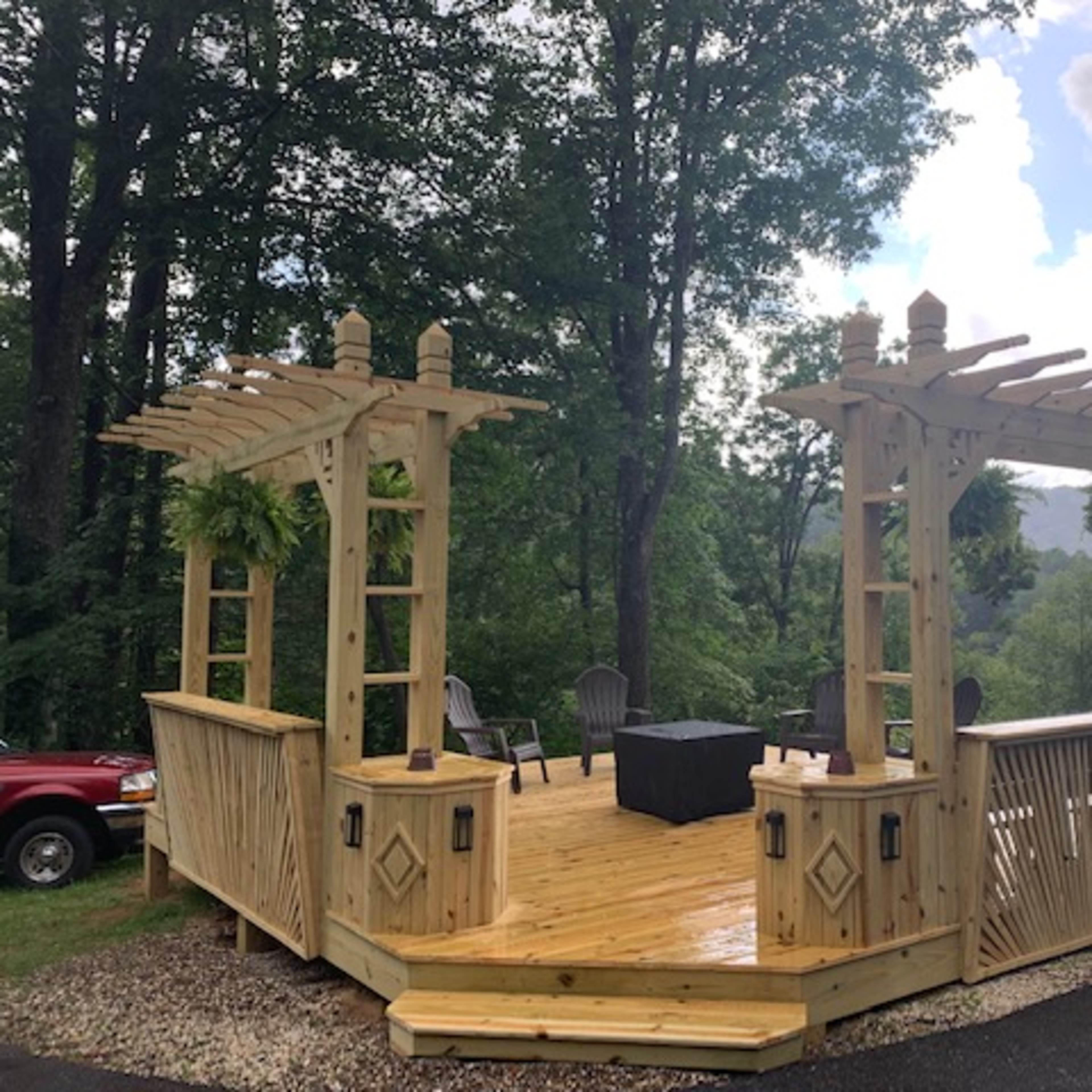 A wooden patio with a pergola design is situated among trees, featuring chairs and a table in a scenic outdoor setting.