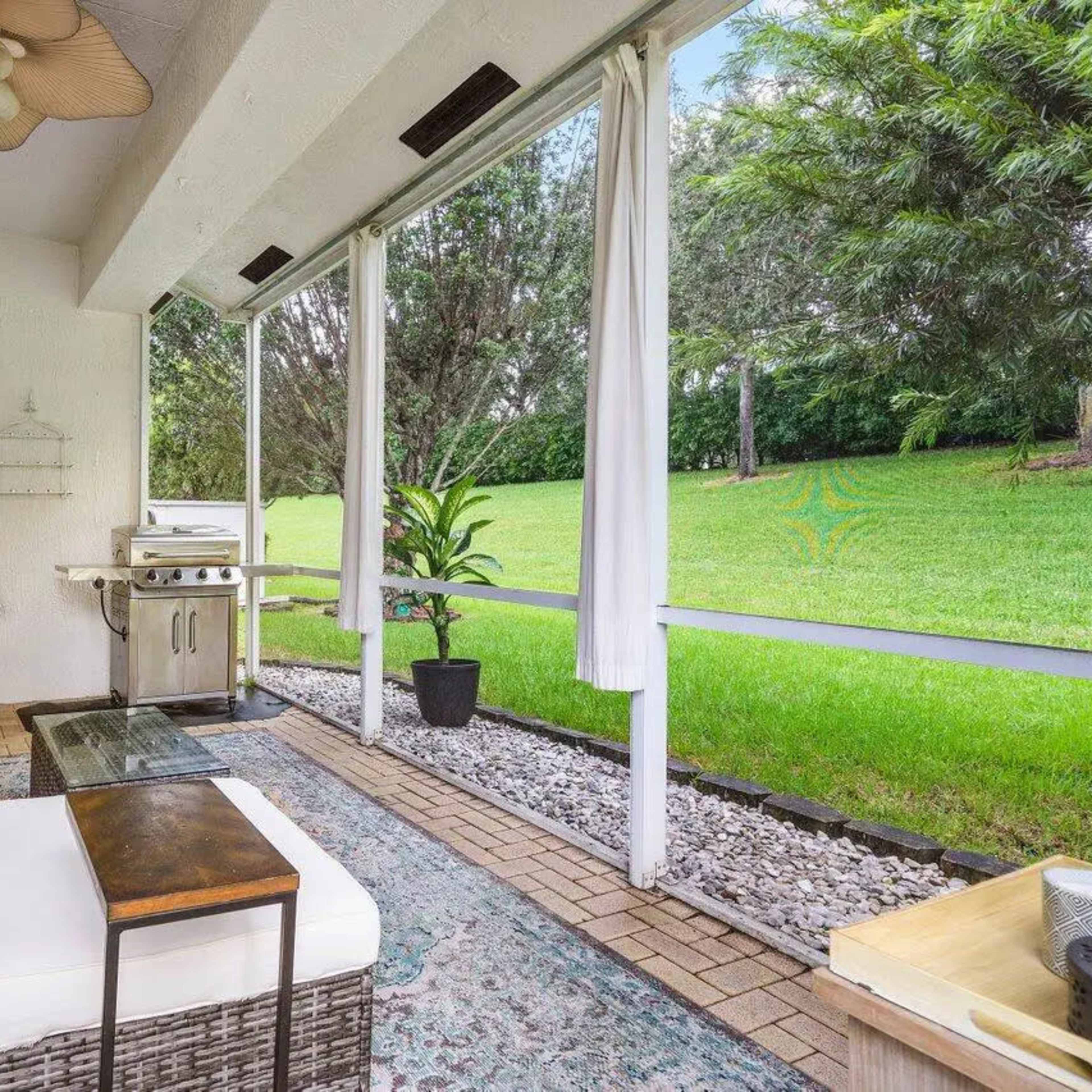 A covered patio area featuring a seating arrangement, a small table, a grill, and a lawn in the background.