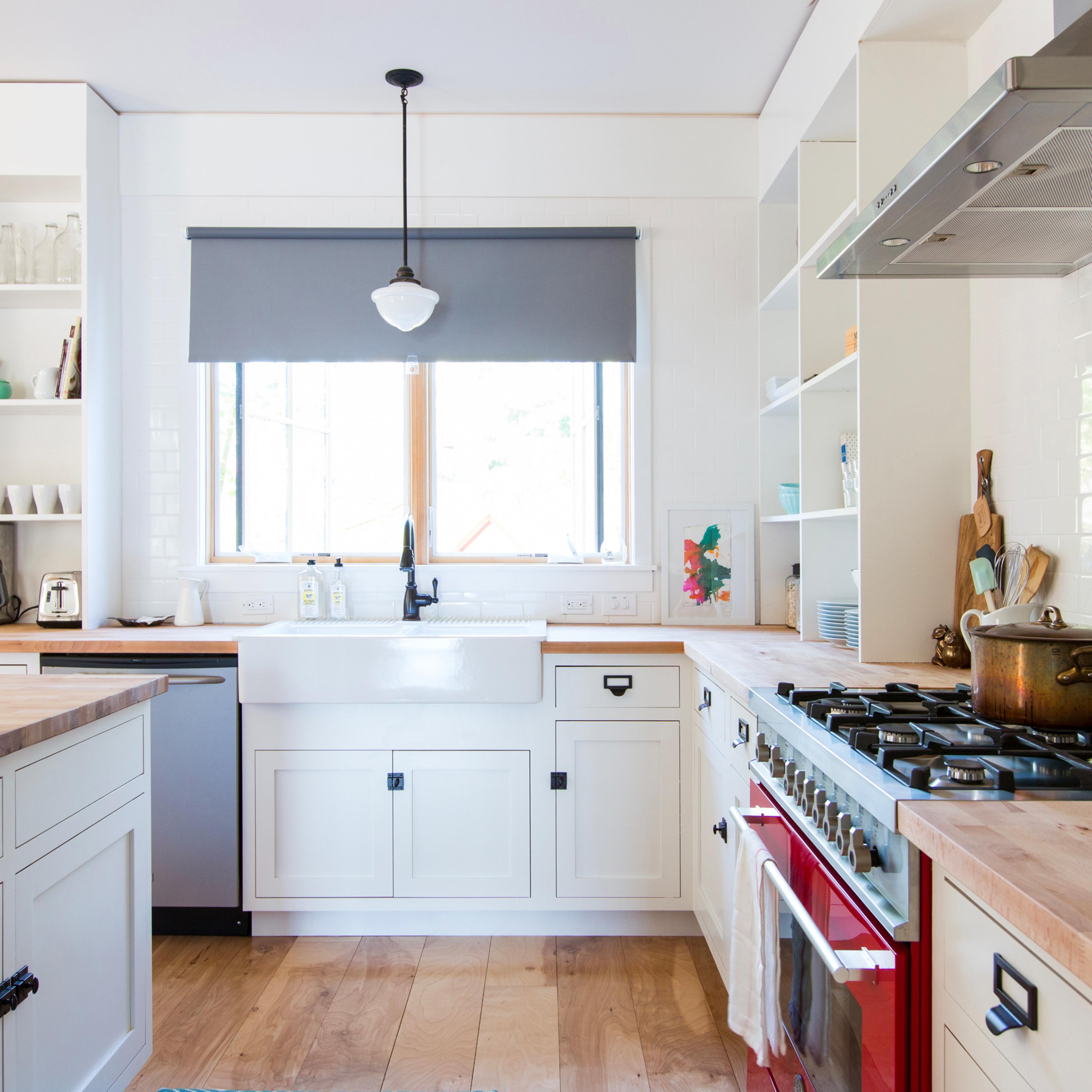 A modern kitchen with a central island, a farmhouse sink under a window, and a red stove next to white cabinetry and open shelves.
