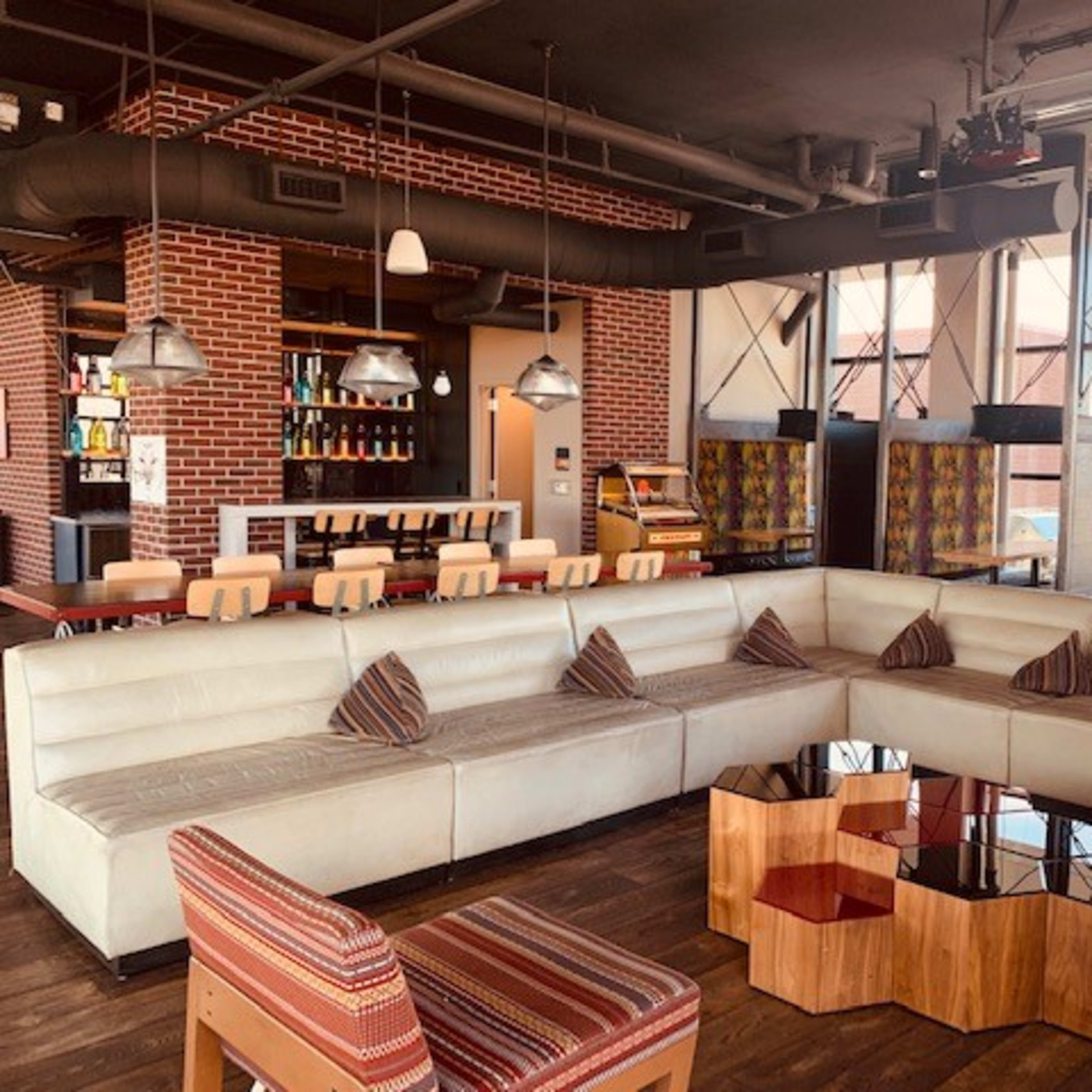 A modern lounge area with a large white sectional sofa, wooden coffee tables, and a bar in the background featuring shelves of bottles.