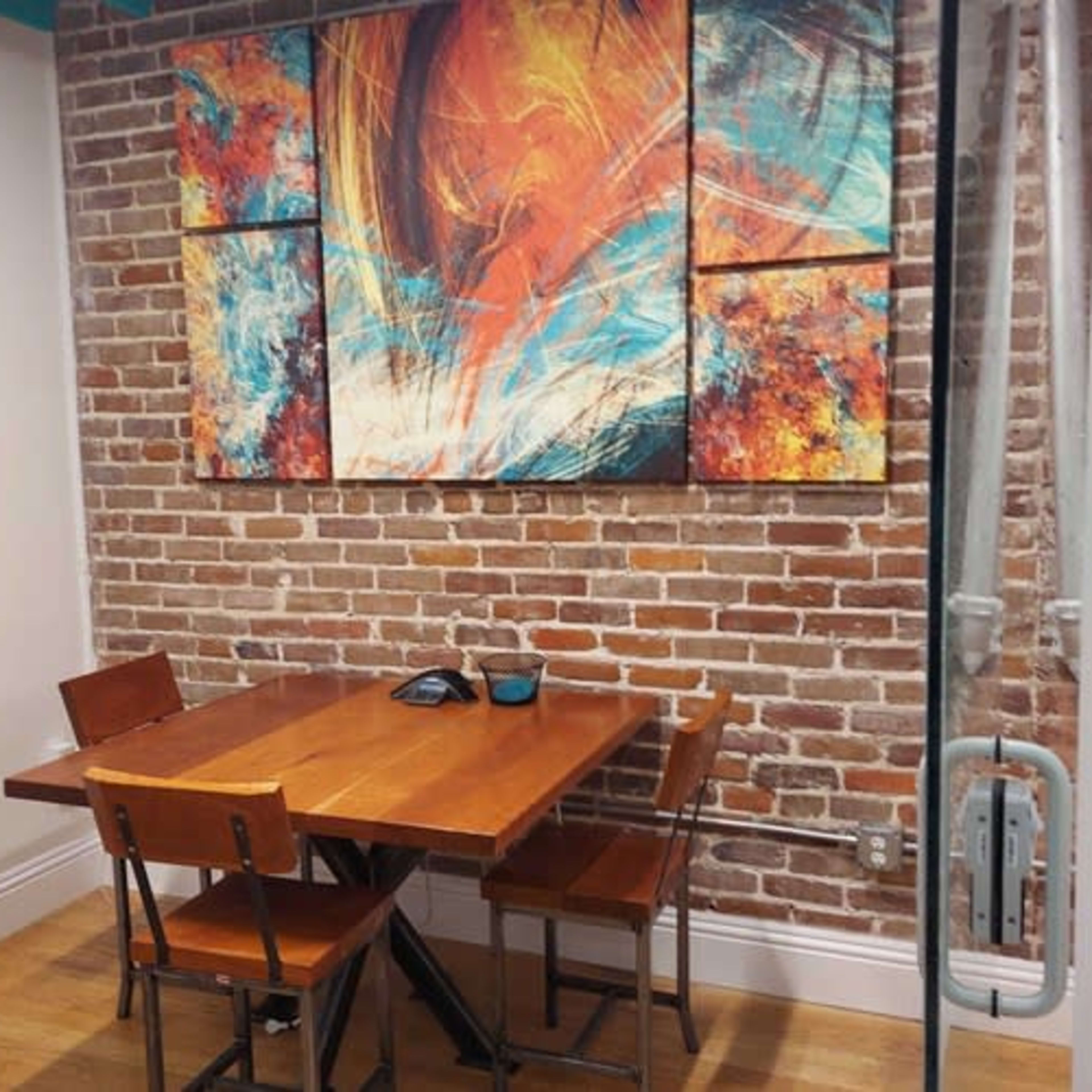A small meeting room with a wooden table and four chairs set against a brick wall, featuring an abstract painting above the table.