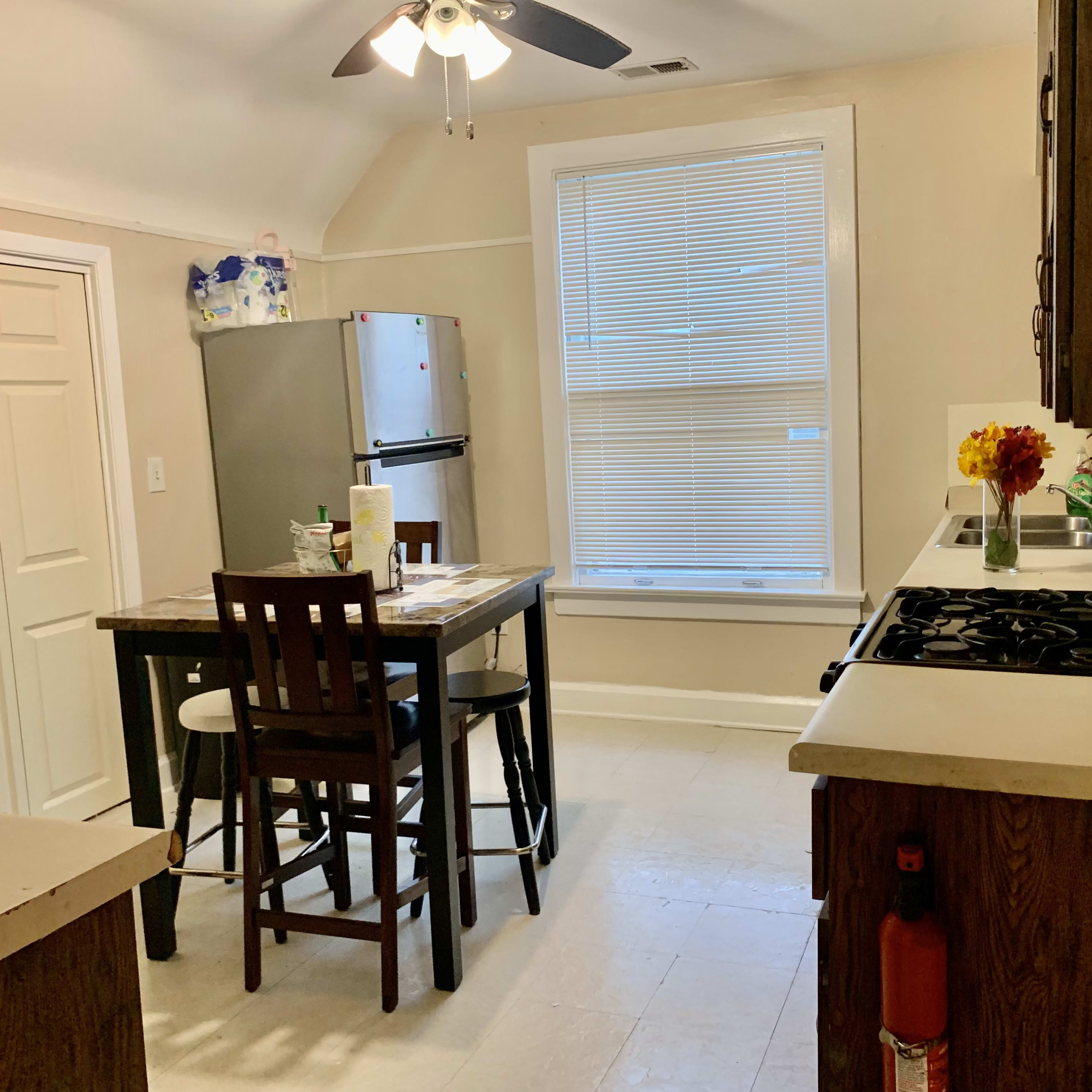 A kitchen features a table with four chairs, a refrigerator, and a window with blinds.
