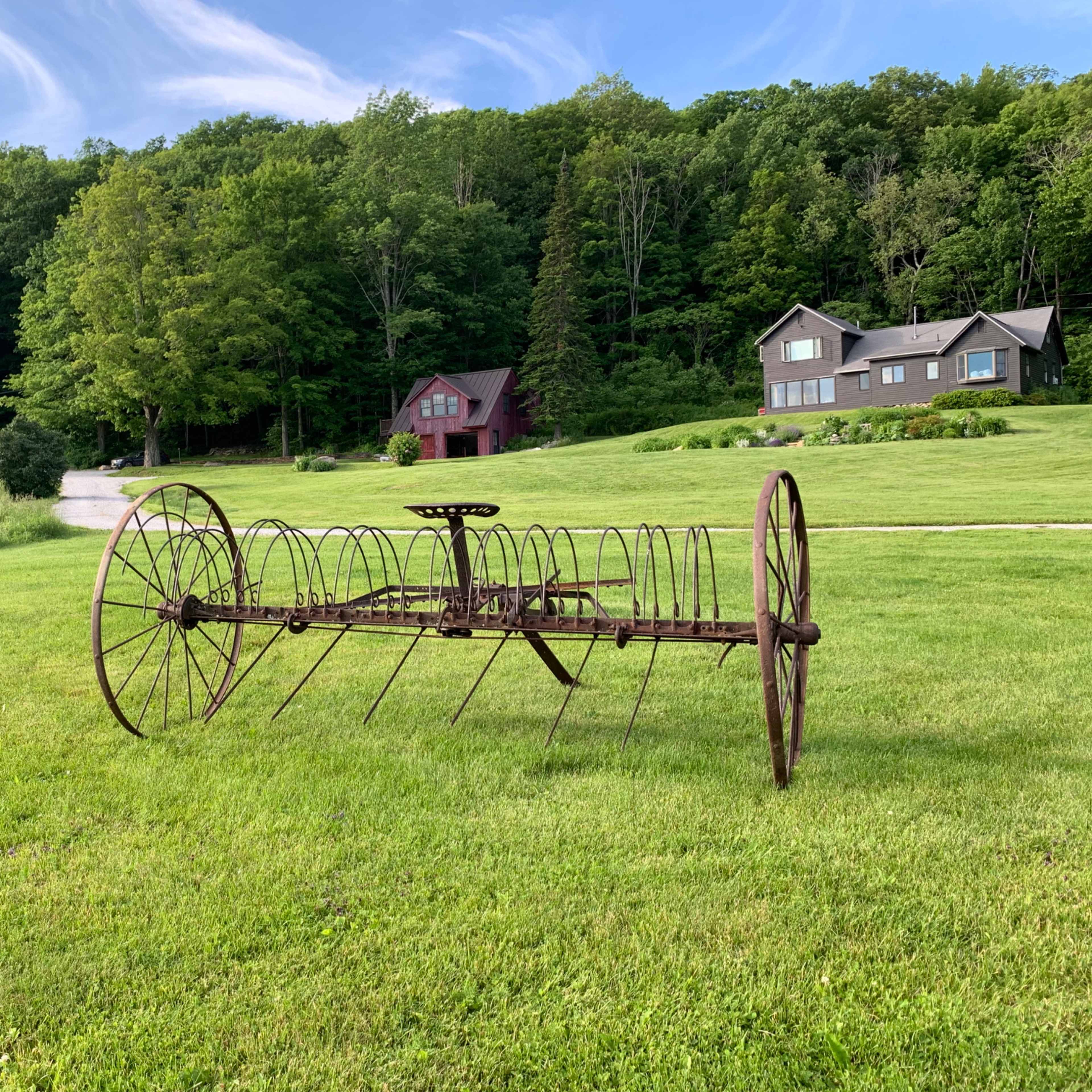 A rusty agricultural rake sits on a grassy field in front of several houses surrounded by trees.