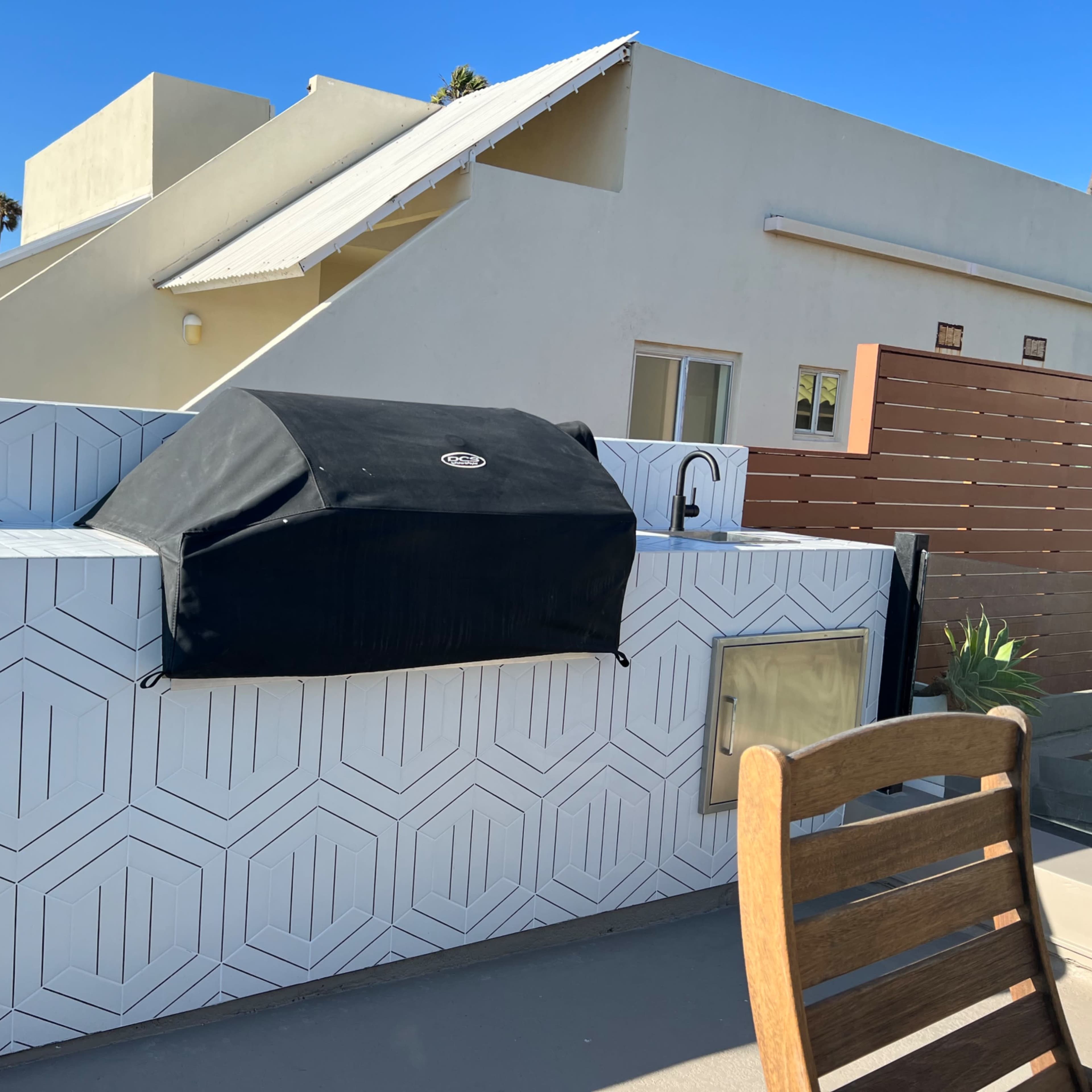 A covered barbecue grill is situated on a geometric-patterned countertop adjacent to a wooden chair and a white building in the background.
