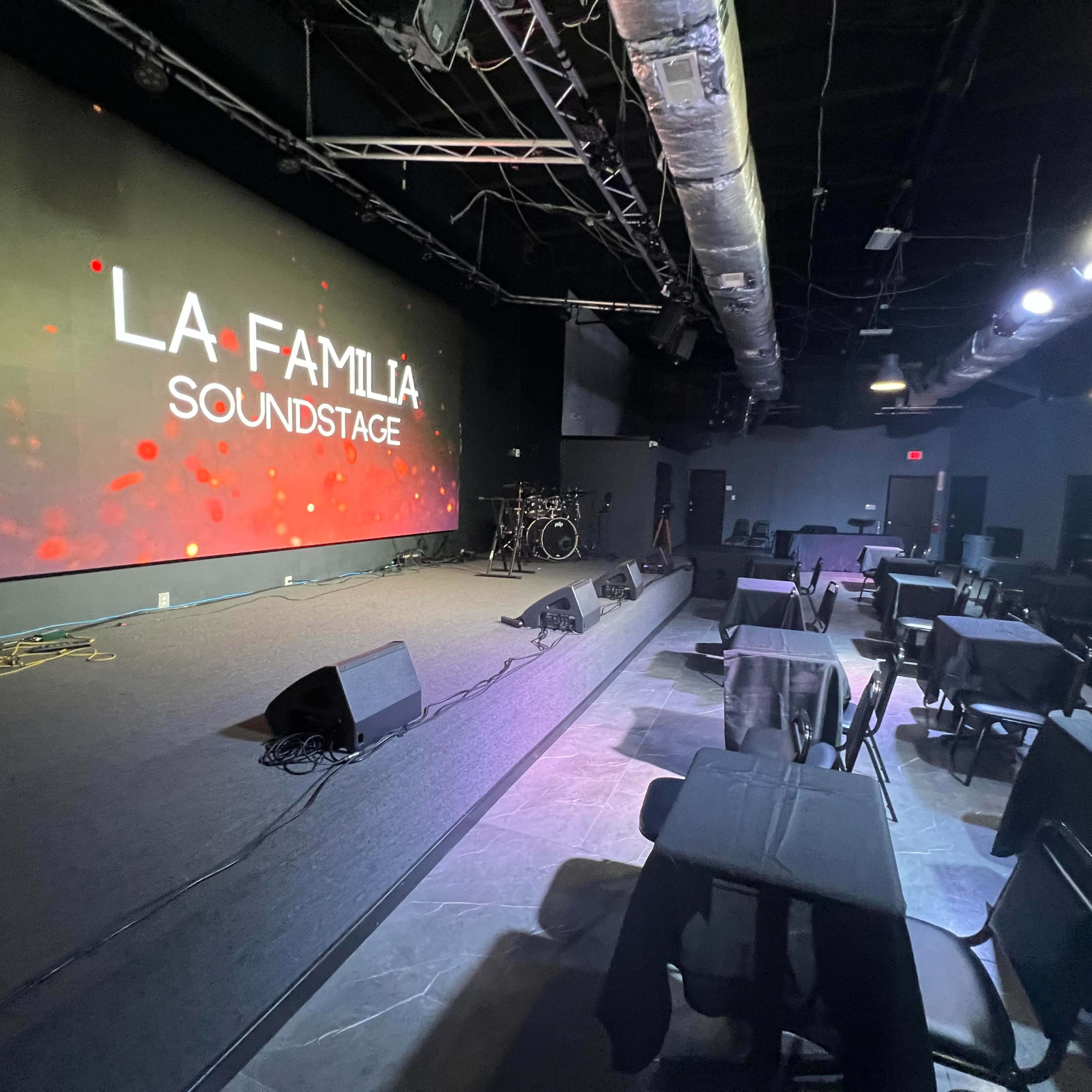 A performance venue with a large screen displaying "LA FAMILIA SOUNDSTAGE," a stage set up with musical instruments, and several black tables and chairs arranged in front.