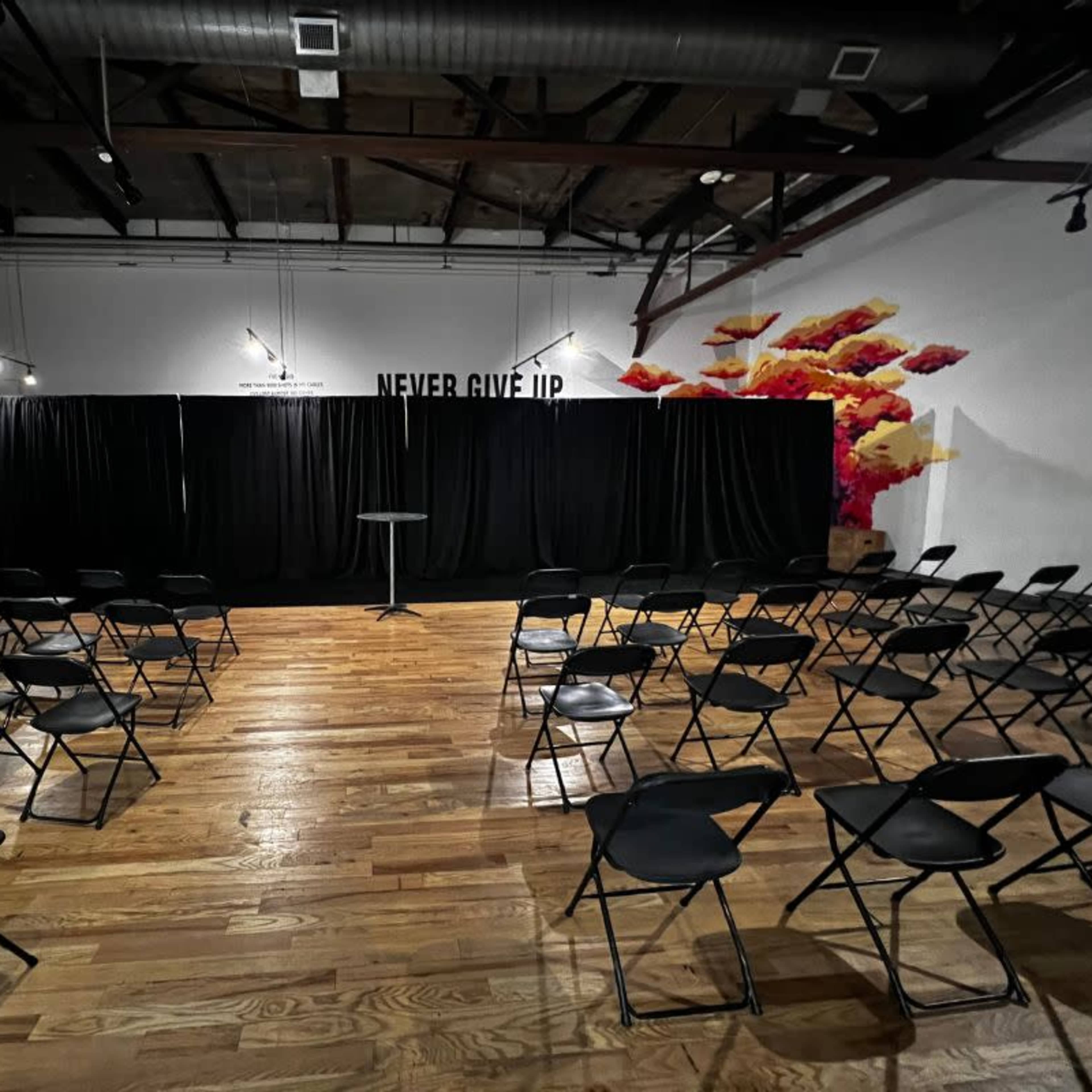 The scene shows a sparsely populated event space with rows of black chairs facing a small stage draped in black curtains, and a wall art featuring the words "NEVER GIVE UP."