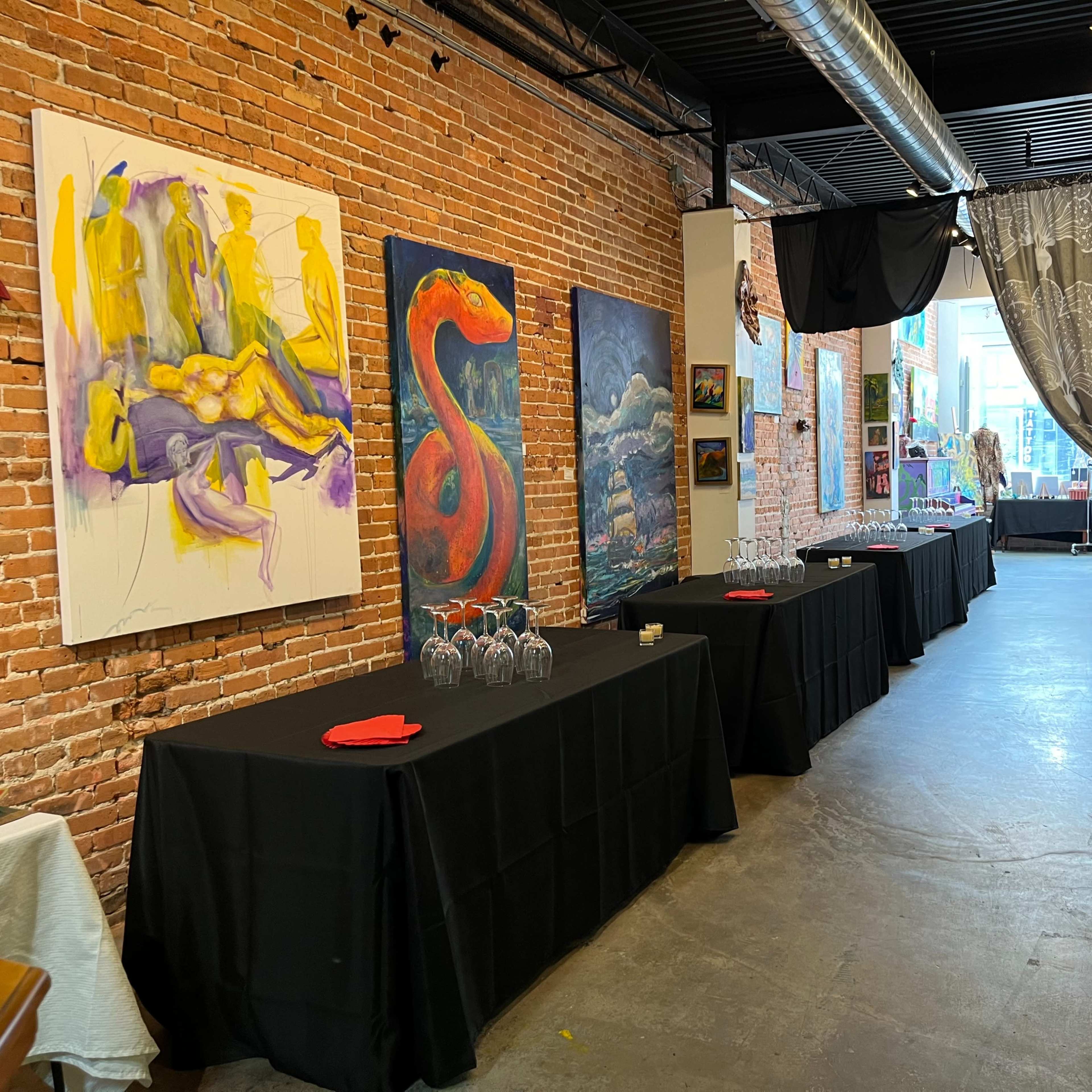 A gallery space featuring exposed brick walls, tables covered with black cloths, and various artworks displayed along the walls.