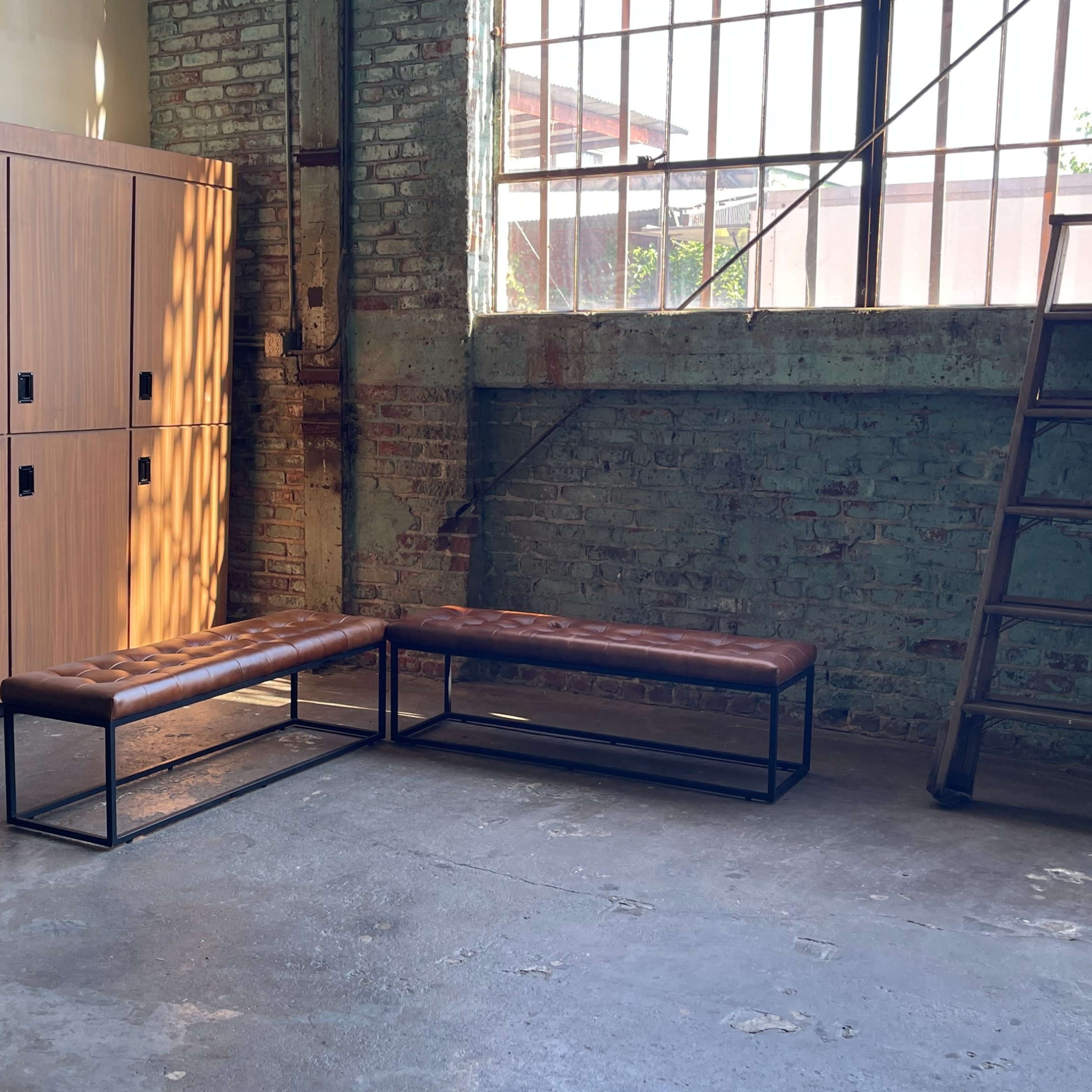 A gym setting with a brown padded bench, a set of dumbbells on the floor, a wooden locker unit, and a ladder against a brick wall with large windows.