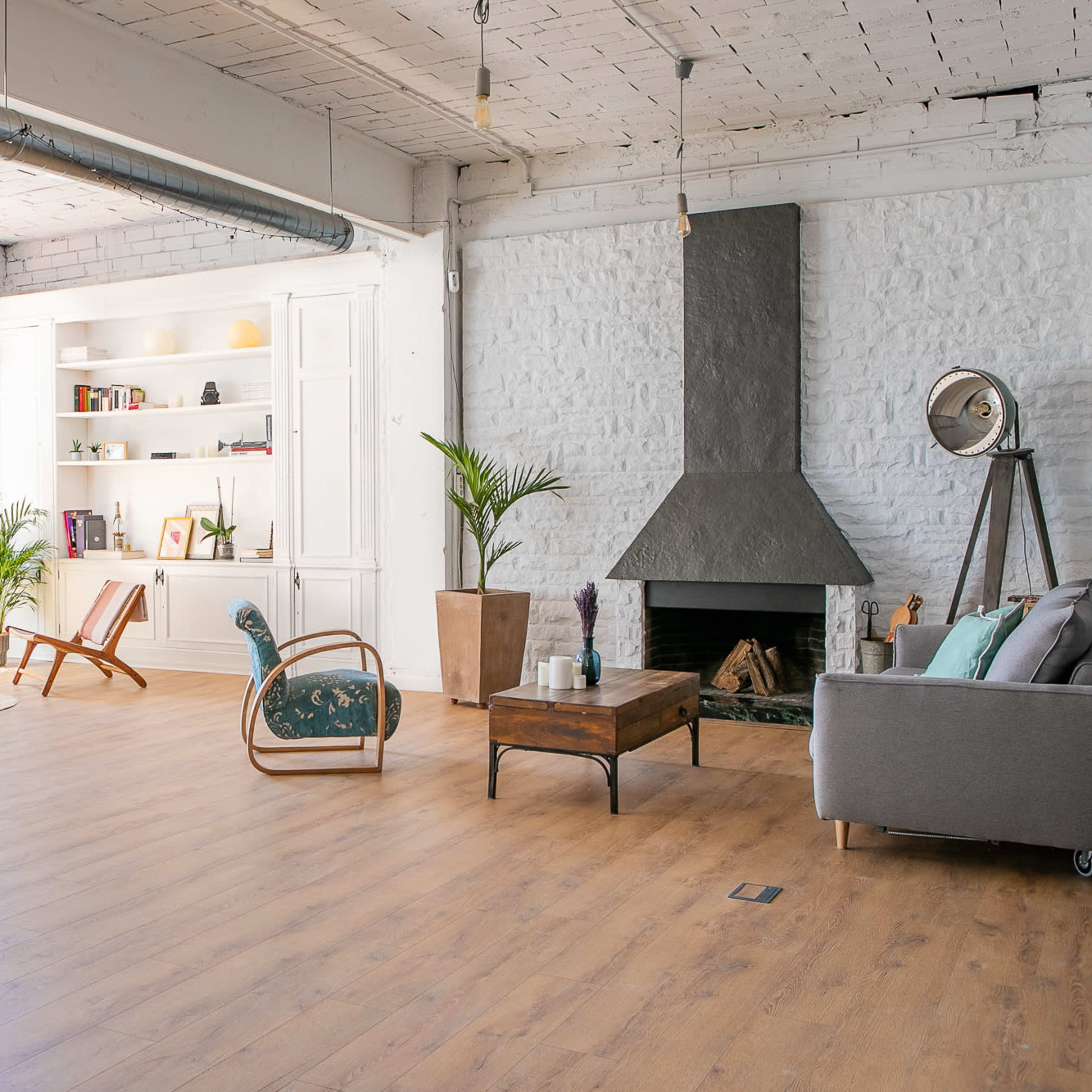 A spacious living area with a large window, a stone fireplace, a wooden floor, and various plants alongside minimalist furniture.