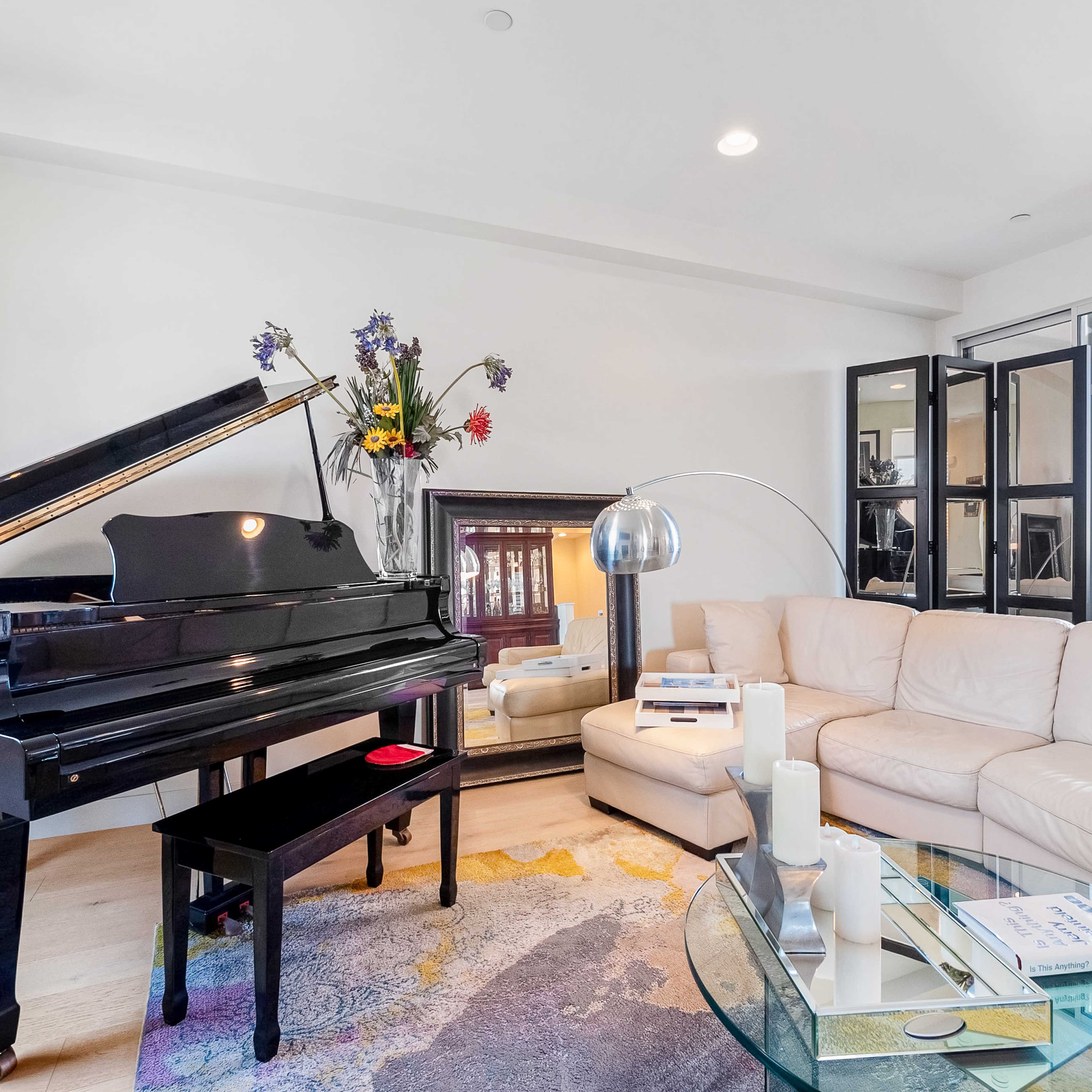 The living room features a grand piano beside a white sectional sofa, a glass coffee table, and large windows with black-frame doors leading outside.