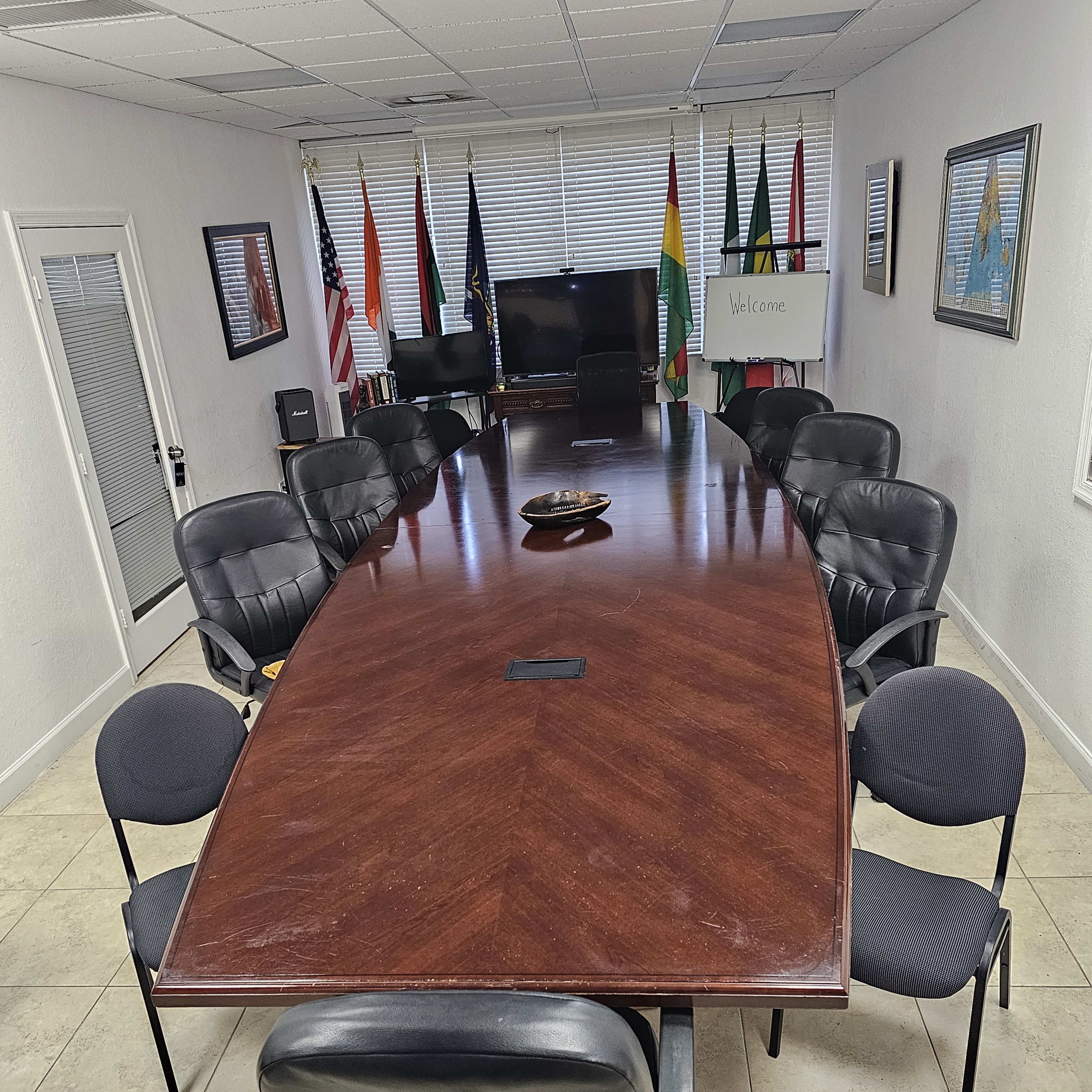 A large, elongated conference table surrounded by black chairs in a well-lit meeting room decorated with flags and framed artwork.
