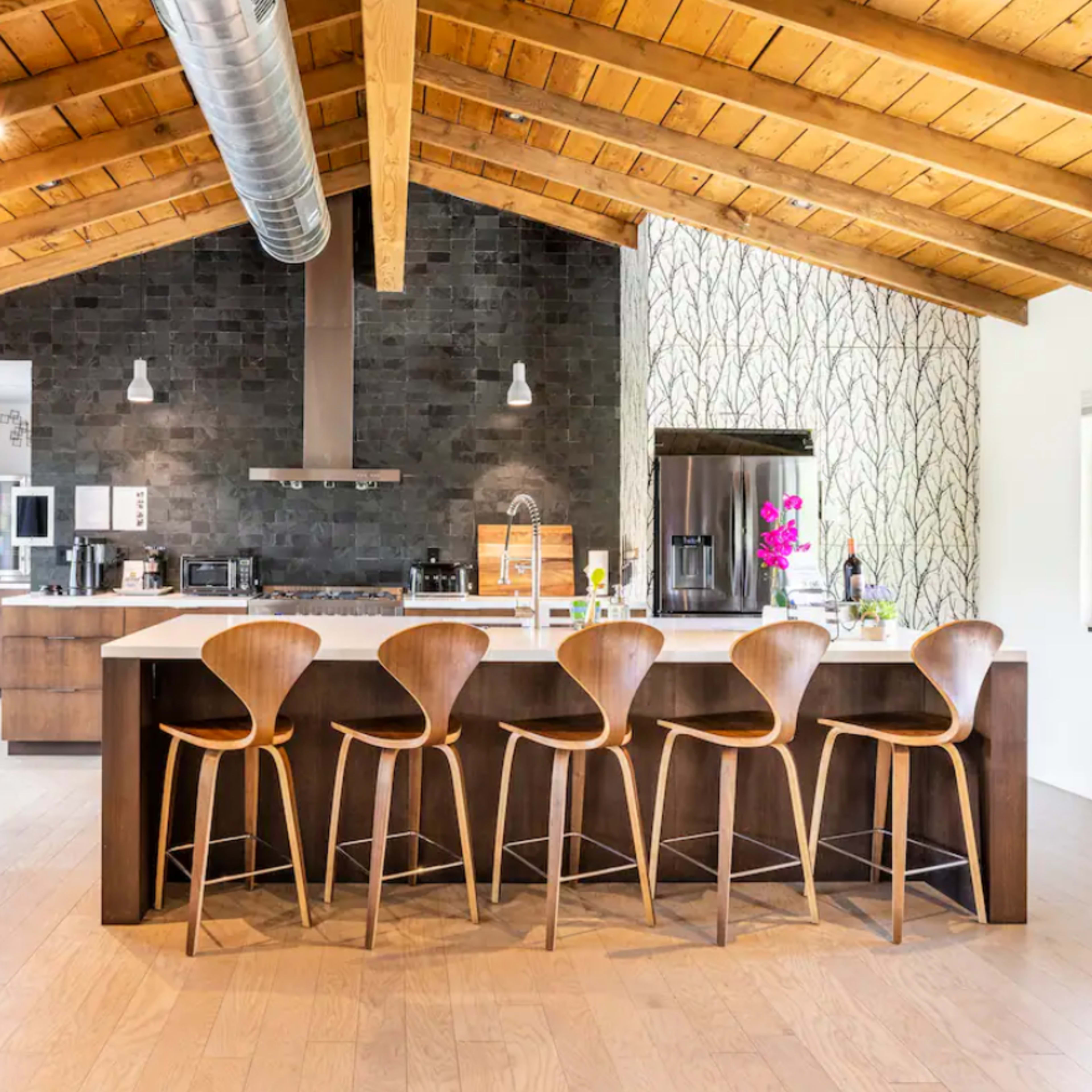 A modern kitchen with a spacious island fitted with wooden bar stools, under a vaulted wooden ceiling and stone accents on the wall.
