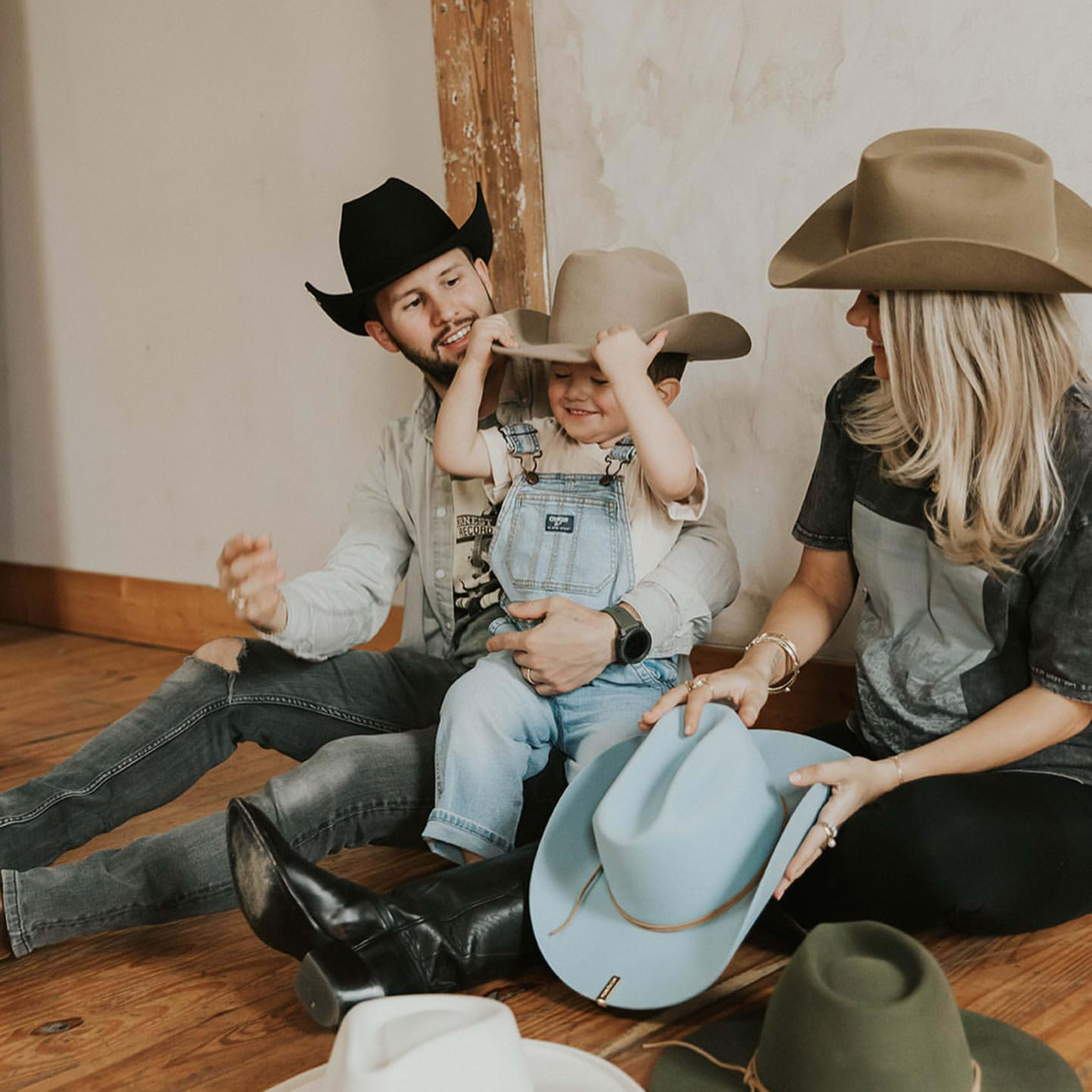 A man and a woman sit on the floor with a child between them, each wearing large hats while surrounded by various hats laid out in front of them.