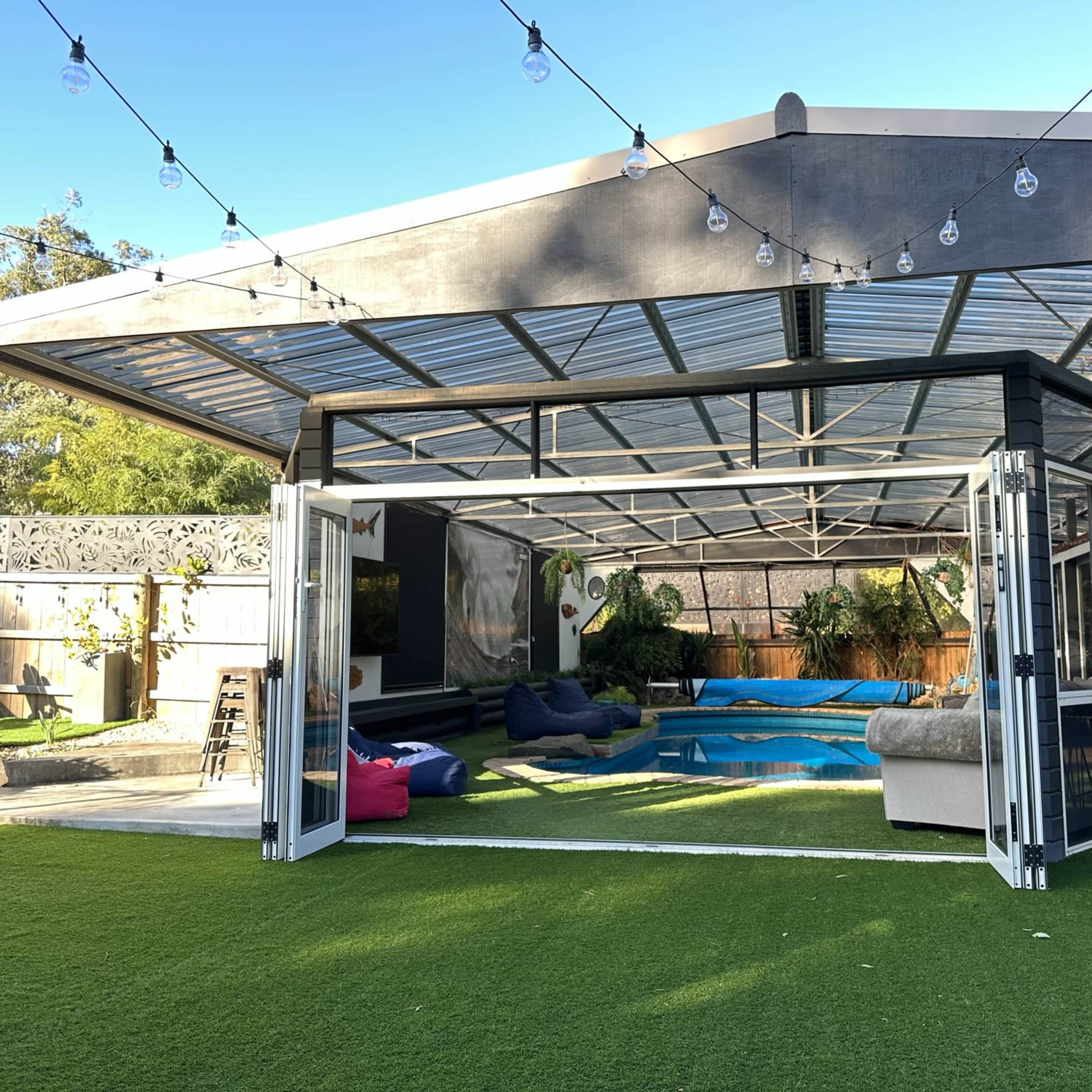 A modern outdoor space features a pool surrounded by greenery and seating, enclosed by glass walls, under string lights.