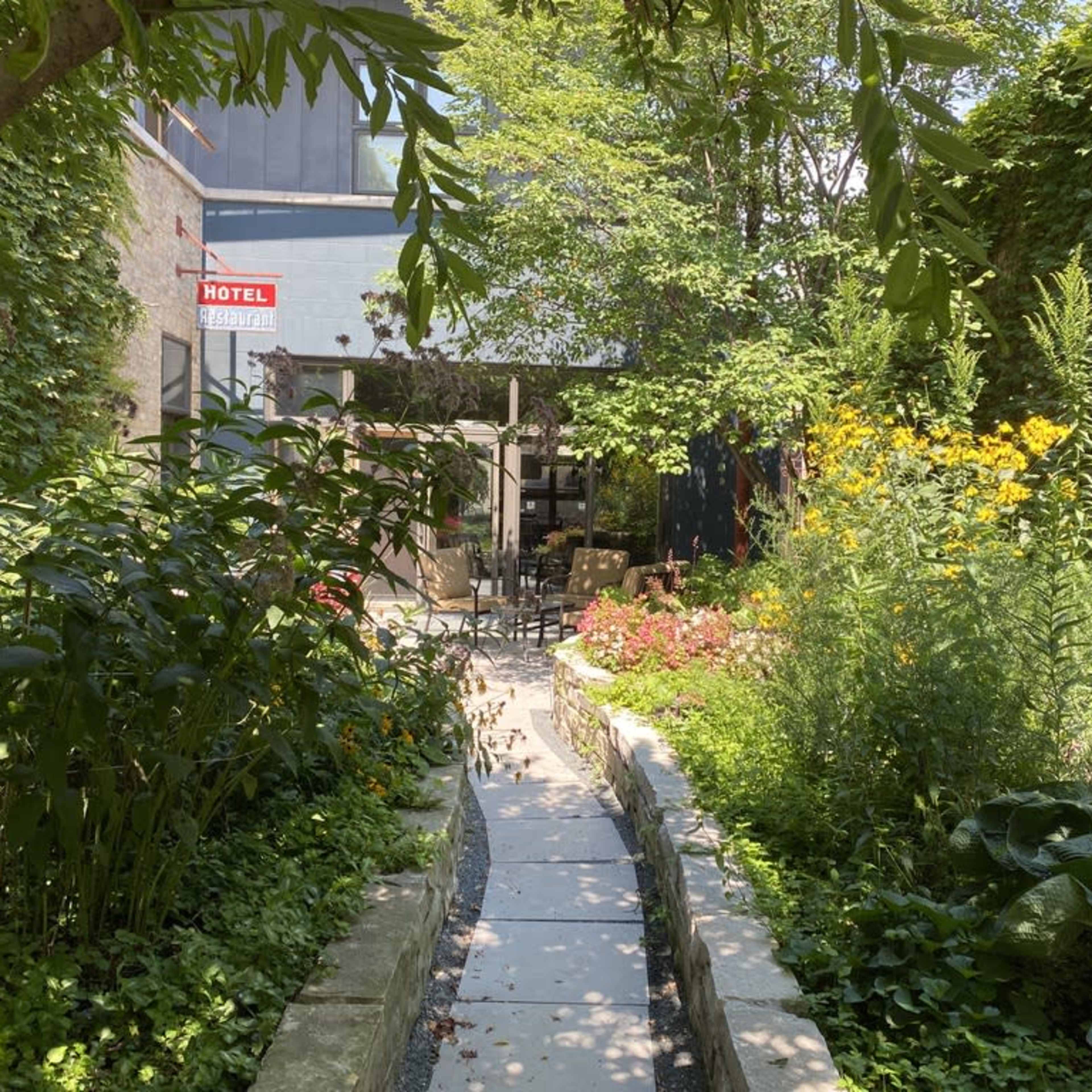 A narrow stone path leads through a lush garden filled with various plants and flowers, with a seating area visible in the background.