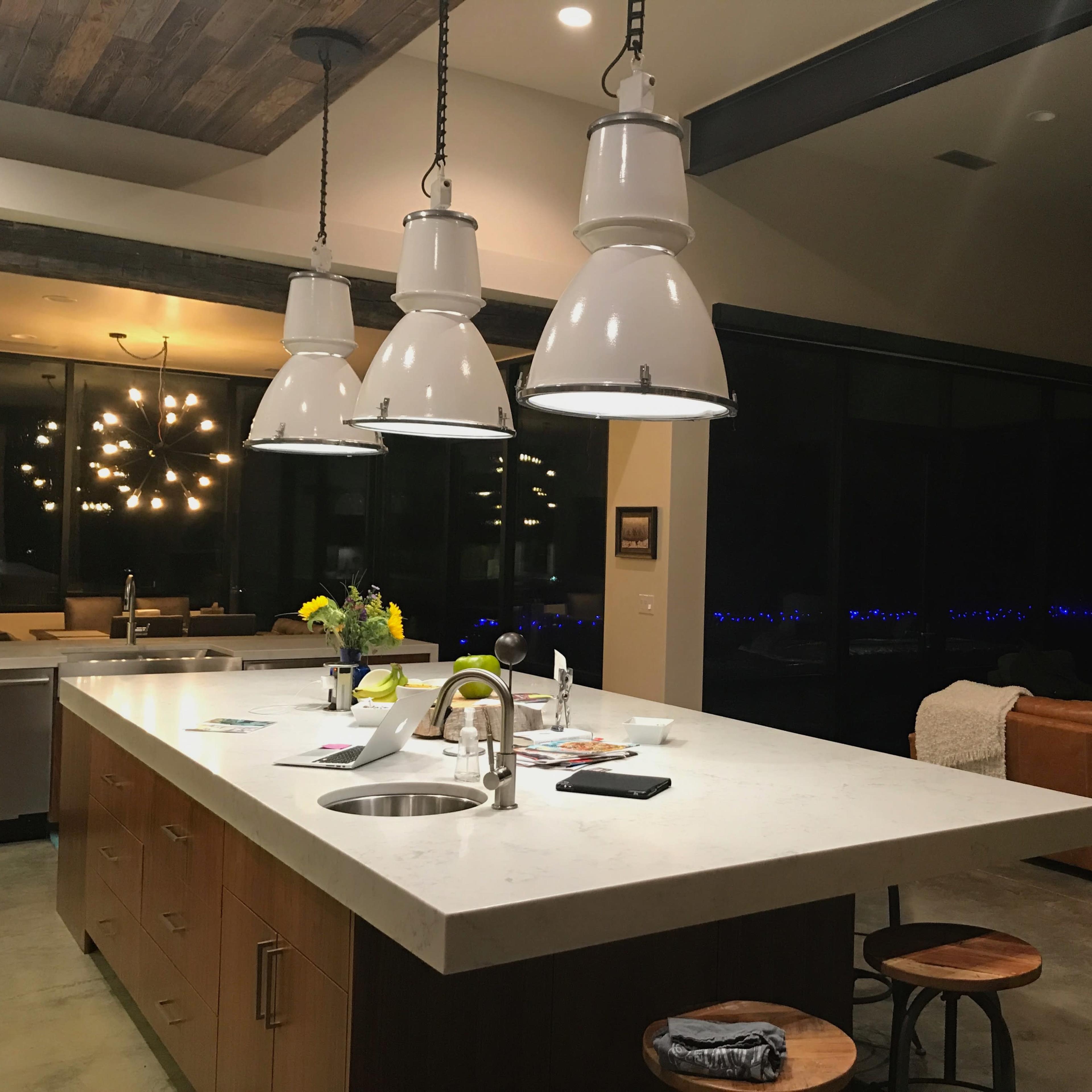 A modern kitchen with a central island, featuring three white pendant lights, wooden cabinets, and a large window revealing a night view.