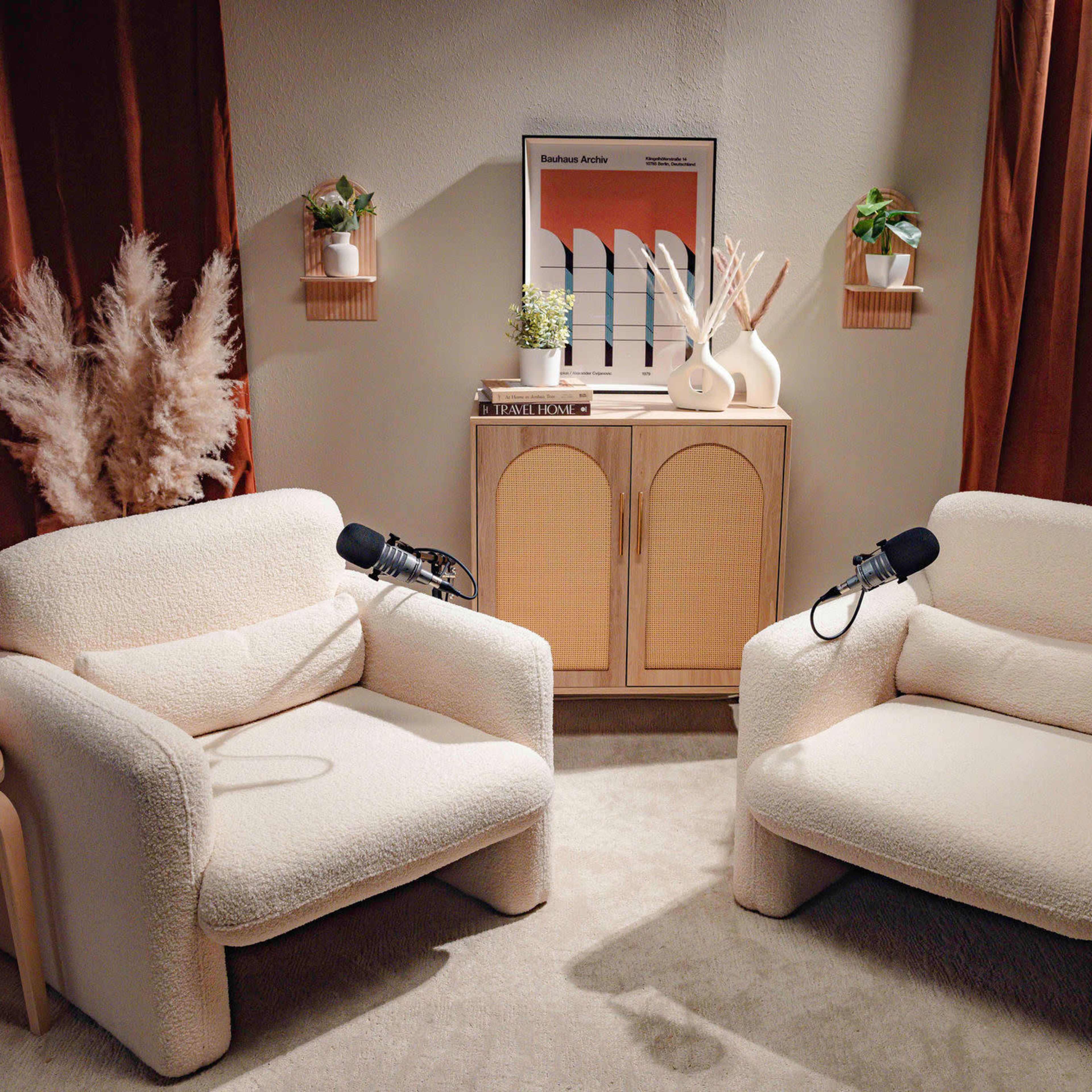 A cozy interior setting with two tufted armchairs, a wooden cabinet in the center, and microphones on each chair, surrounded by plants and decorative elements.