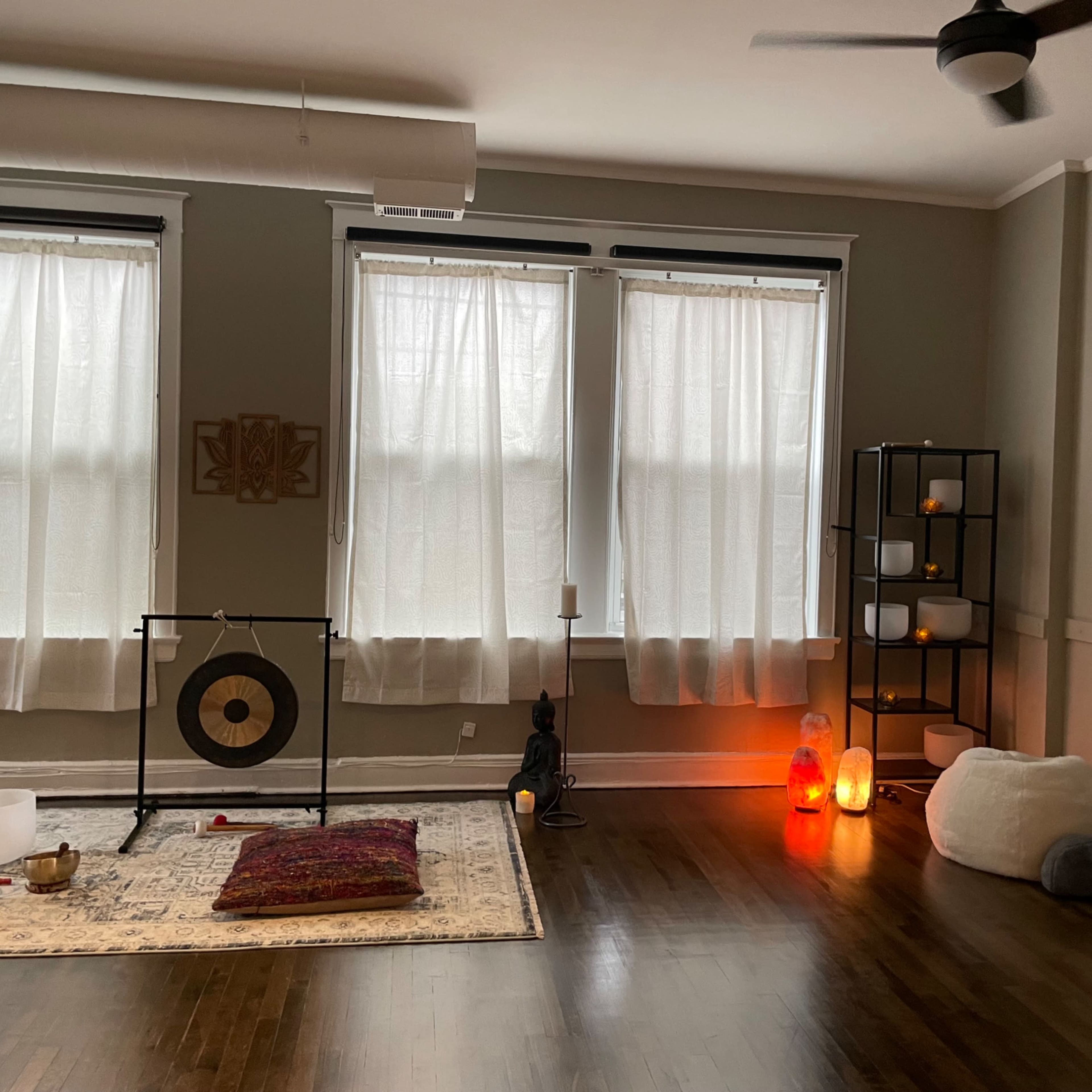 The room features three large windows with sheer curtains, a gong on a stand, various candles and lamps, a wall shelf with decorative items, and a soft seating area with floor cushions.