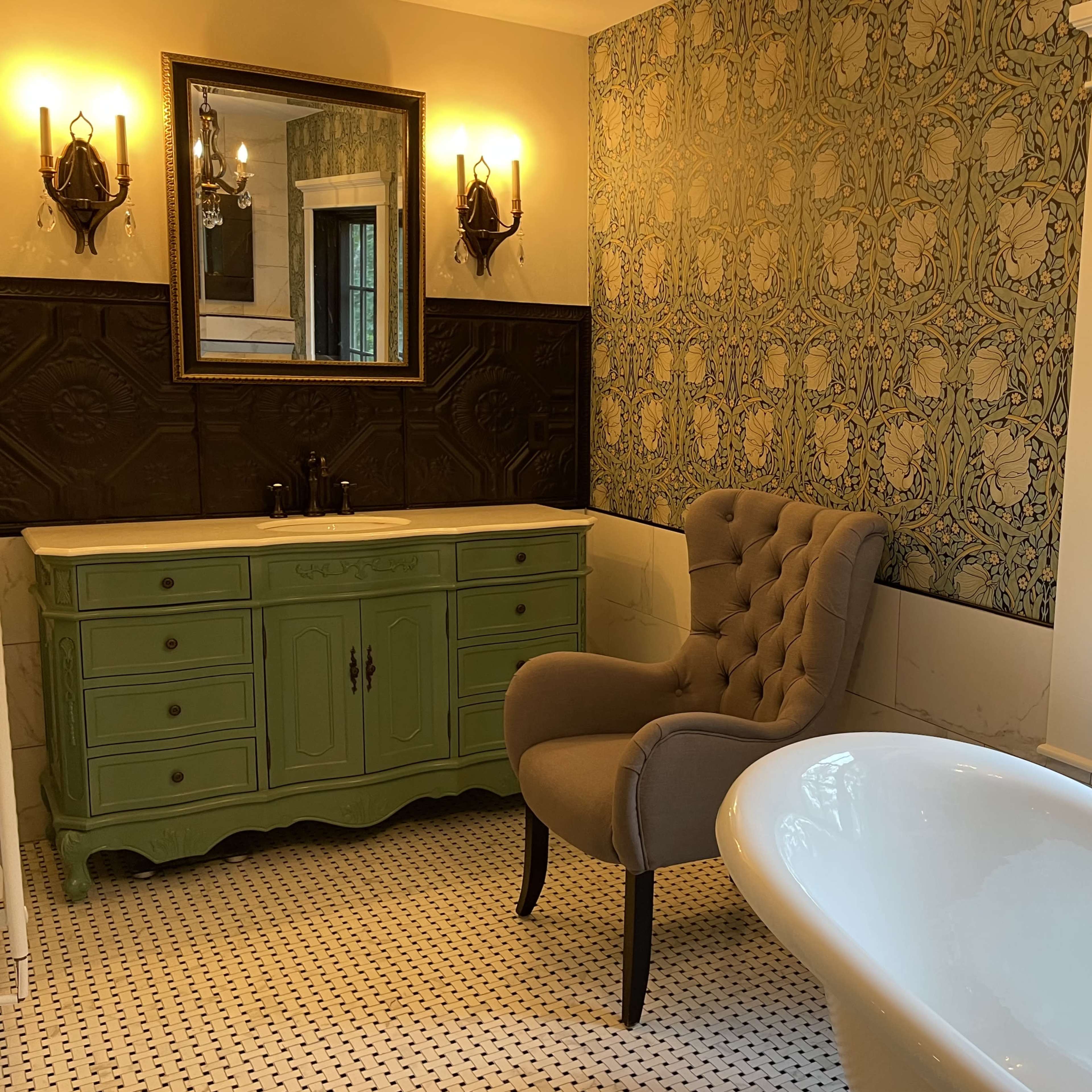 A bathroom features a green vanity, a tufted gray armchair, a freestanding white bathtub, and large windows with a view of the outdoors.