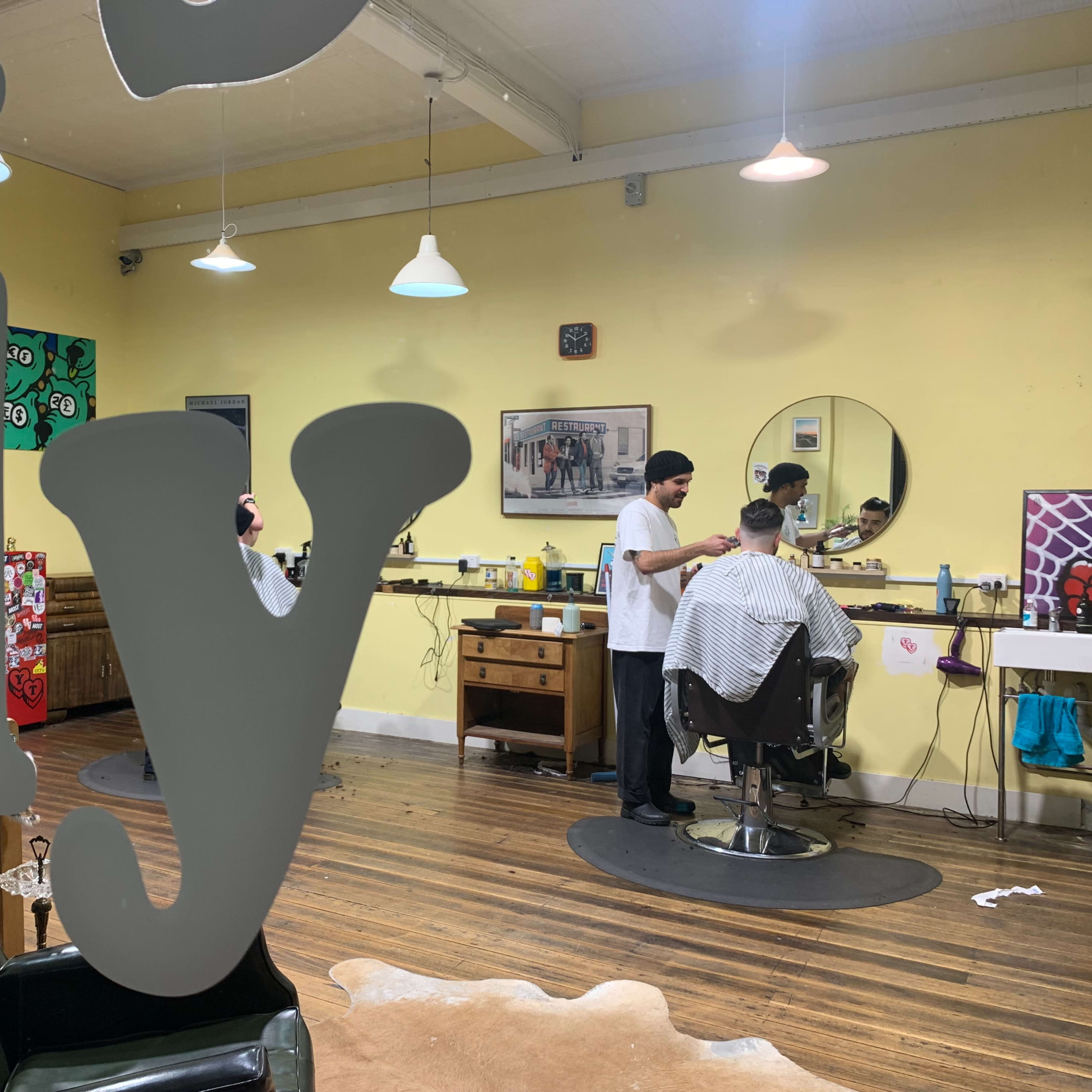 A barbershop interior with two barbers attending to clients in styling chairs, surrounded by mirrors and colorful artwork on the walls.