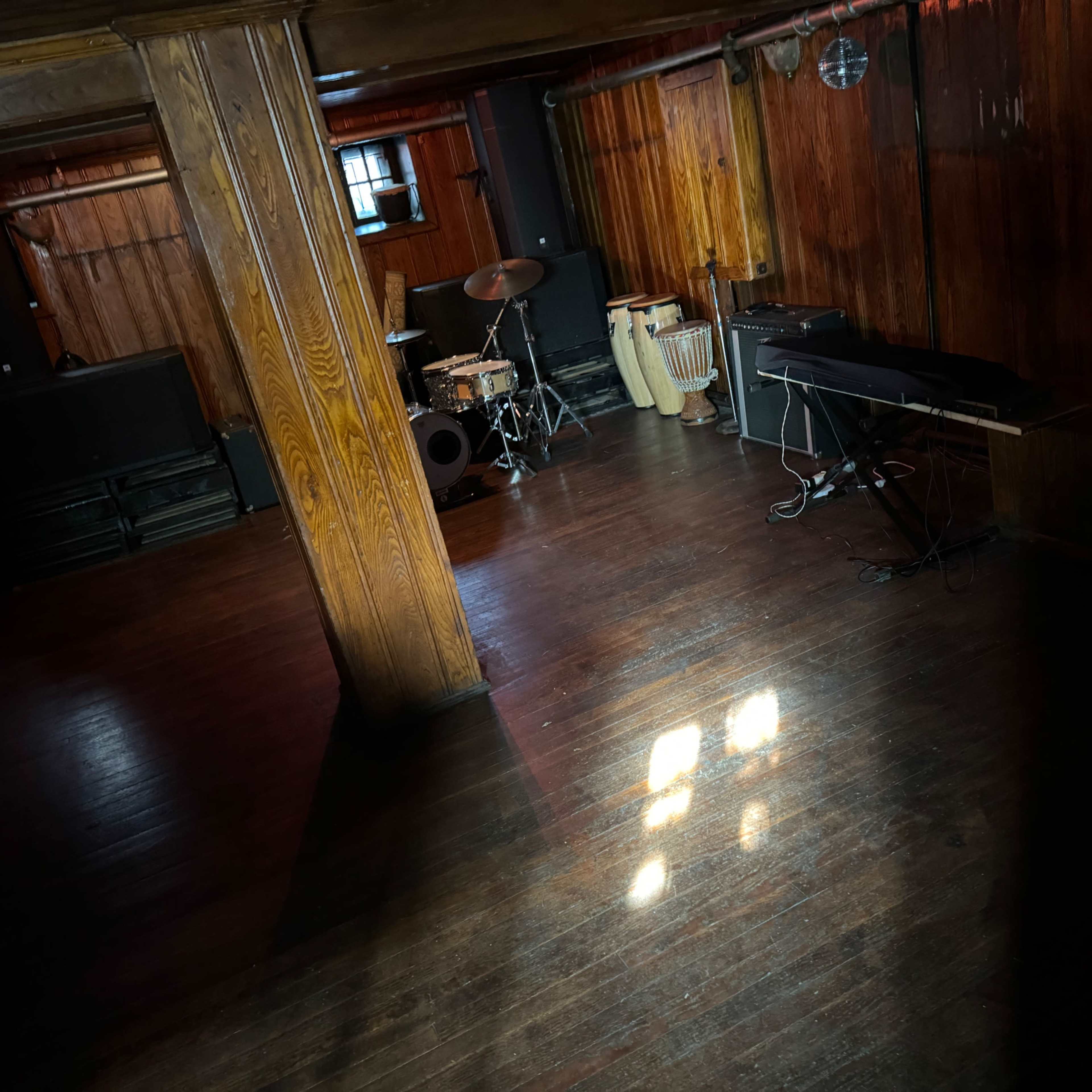 A wooden-floored room with musical instruments, including a drum set and a keyboard, positioned under low lighting.