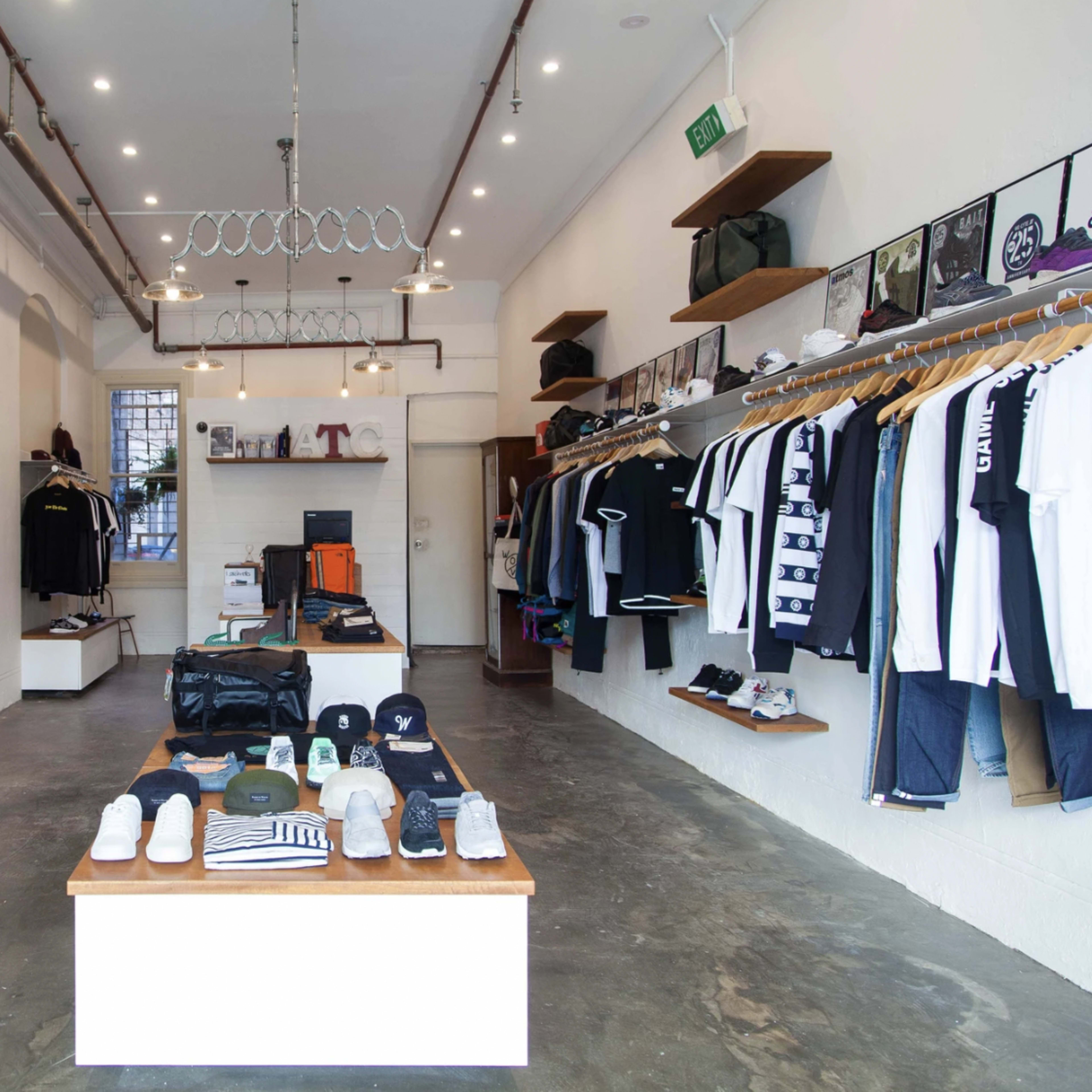 A clothing store interior with shelves displaying various garments, organized neatly along the walls, and a central table featuring shoes and accessories.