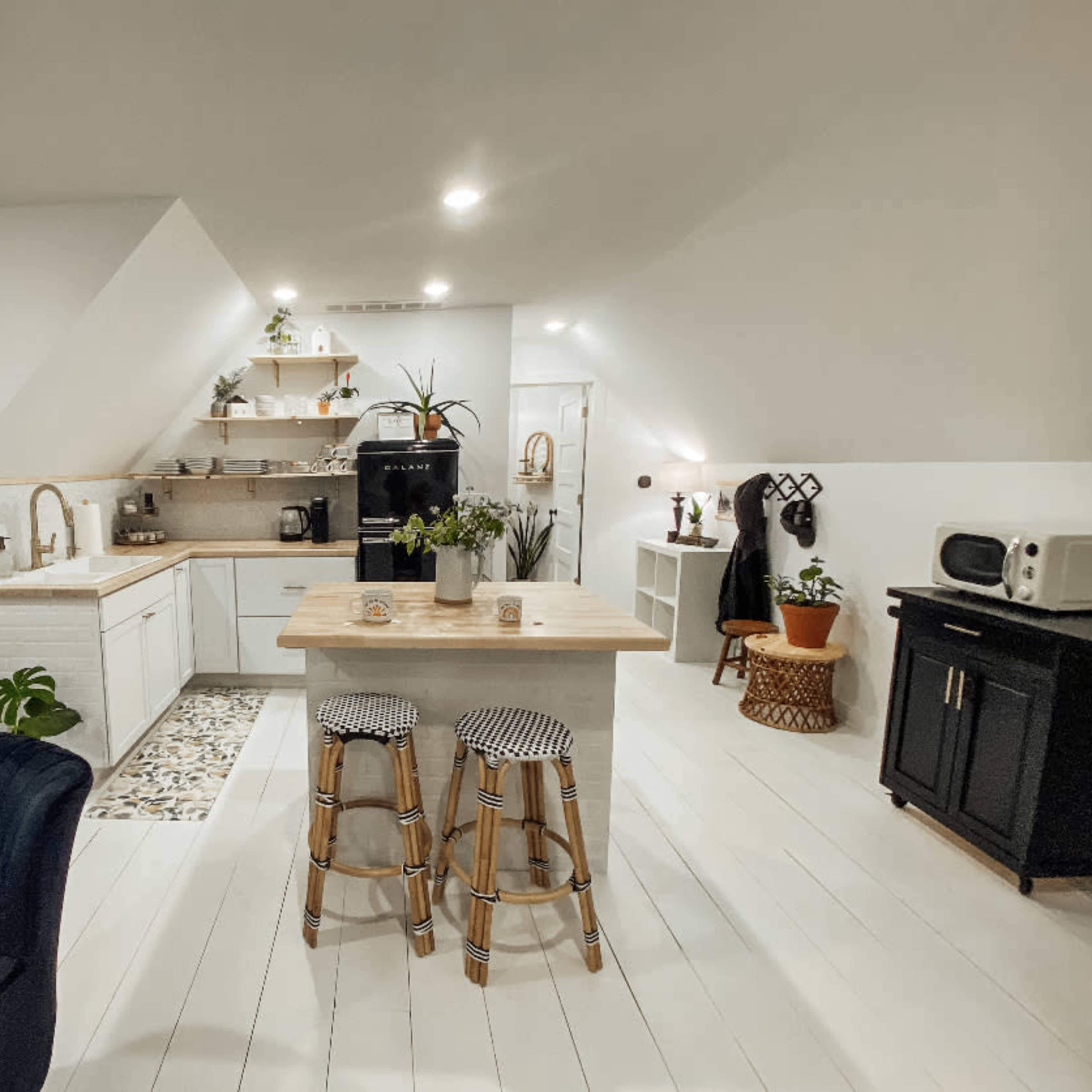 A modern, minimalist kitchen and living area with white walls, a wooden island, and two bar stools, alongside a small workspace featuring a desk and chair.