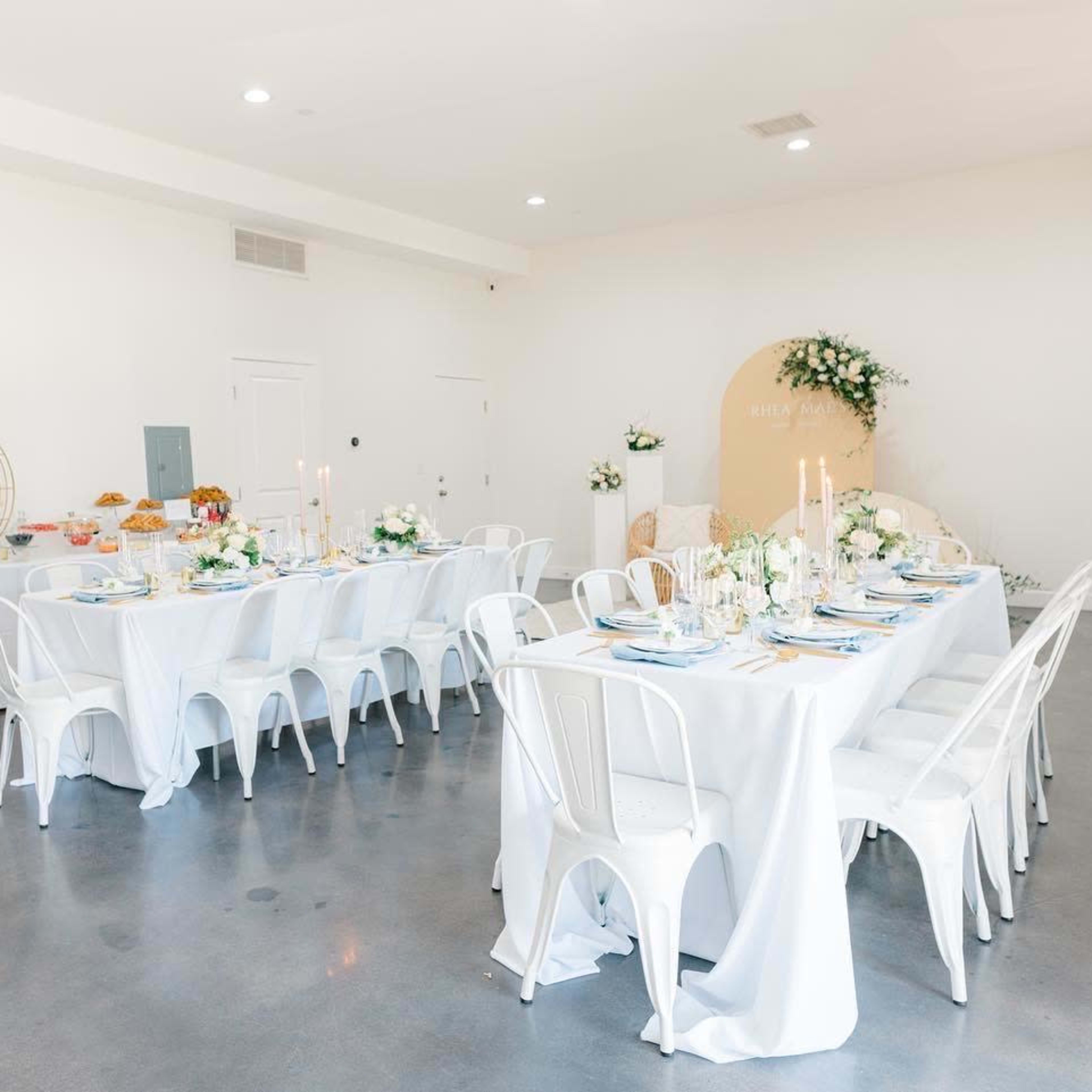 A light-filled event space with two long tables covered in white tablecloths, decorated with floral arrangements and candles.