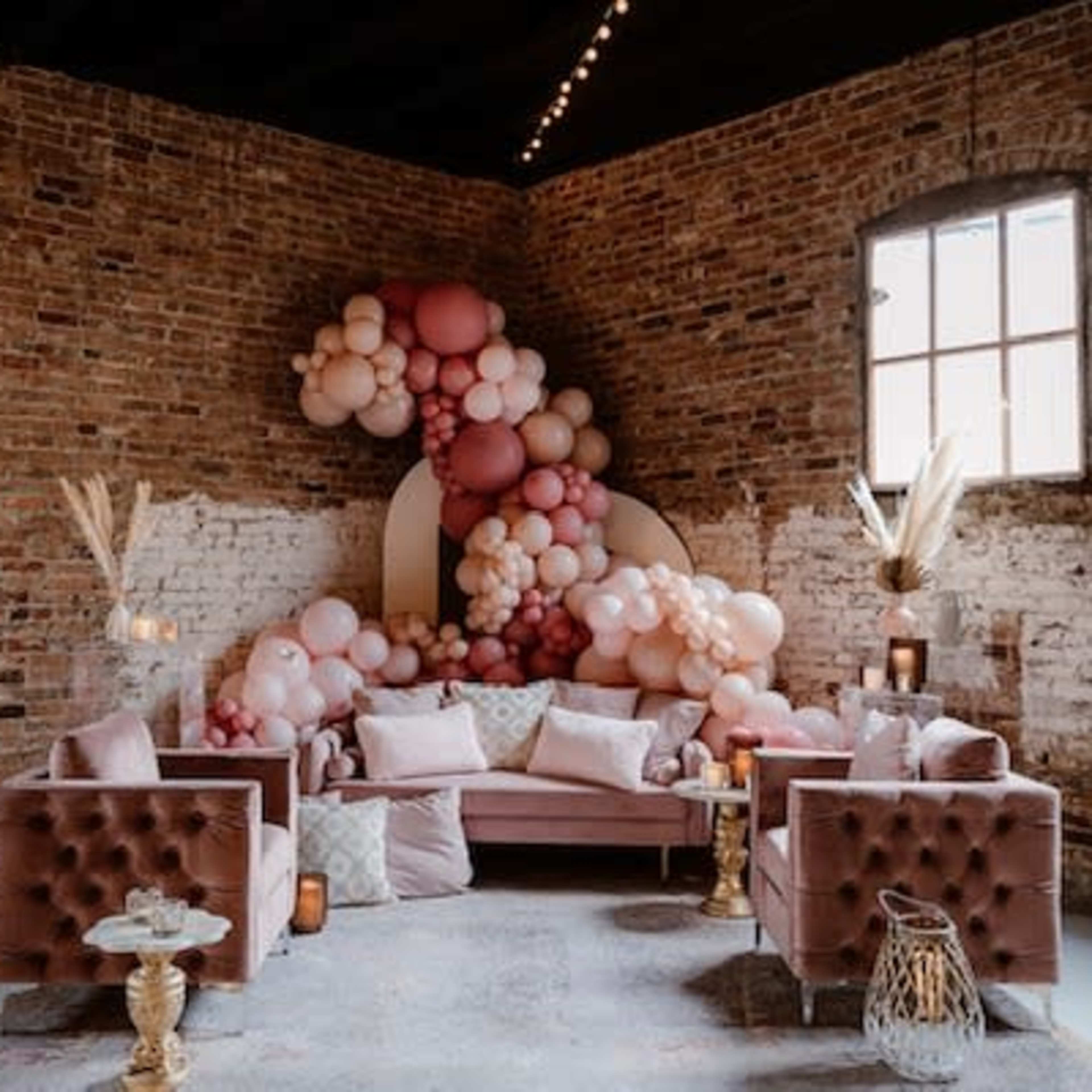 A cozy lounge area features a pink balloon arrangement against a brick wall, with a plush sofa and chairs arranged around a decorative table.