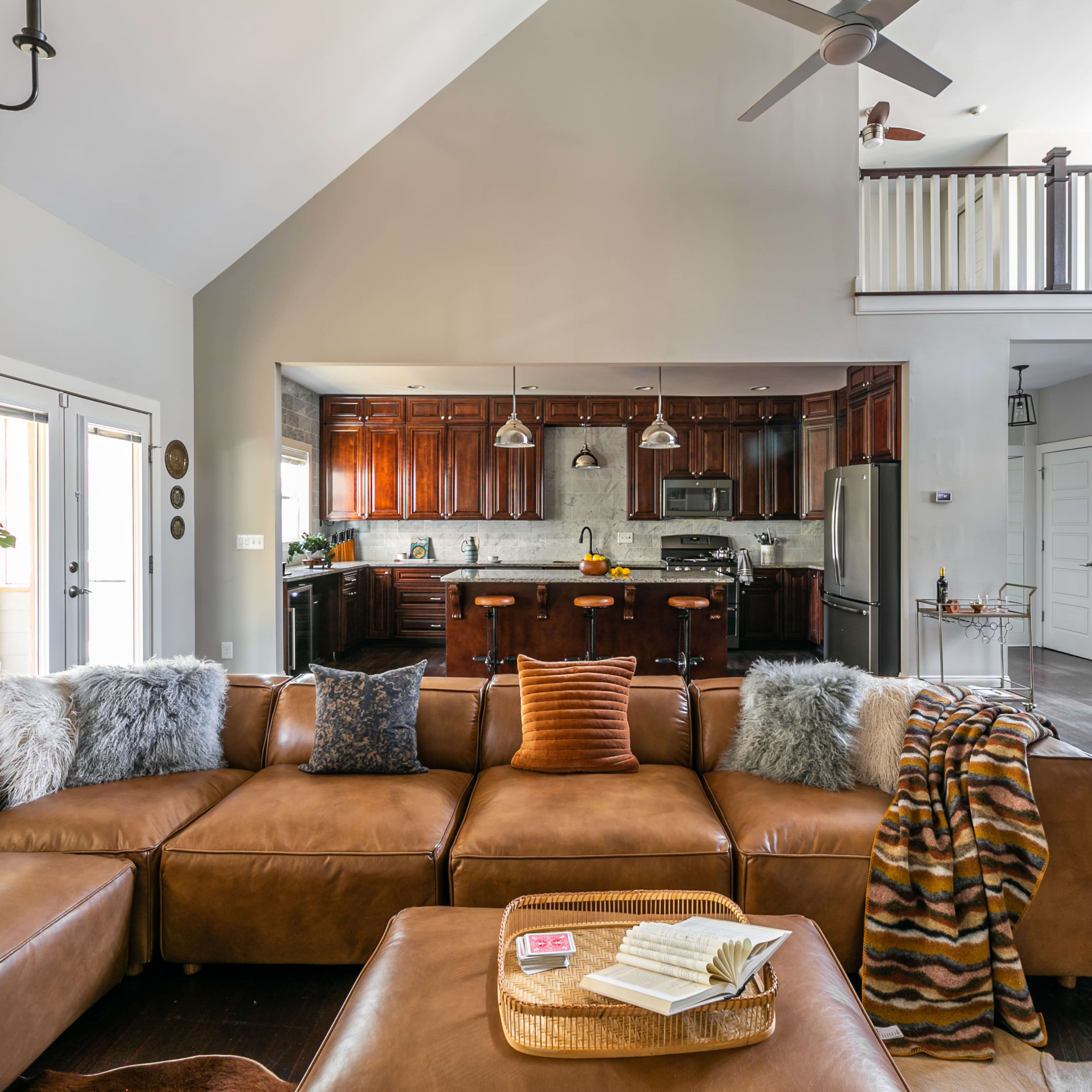 A spacious living room features a large brown leather sectional sofa, an open kitchen with dark wood cabinetry, and a staircase complemented by white railings.